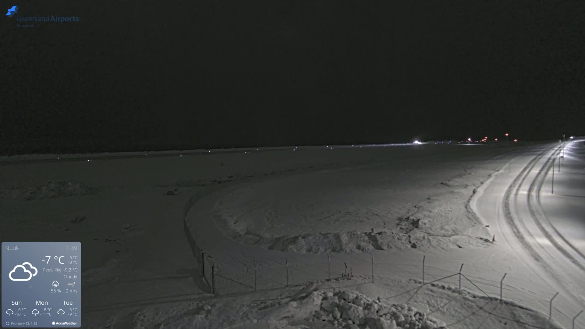 A snow-covered landscape at night, illuminated by lights, shows a road curving through the scene towards a dimly lit horizon under a cloudy sky.