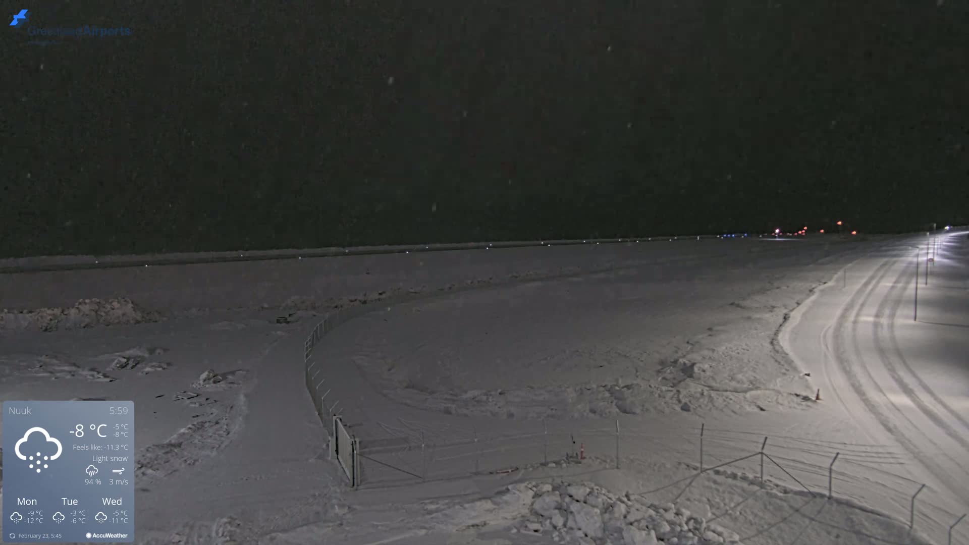 A snow-covered landscape at night under light snowfall, showing a road with tire tracks and a fence enclosing a snowy area.
