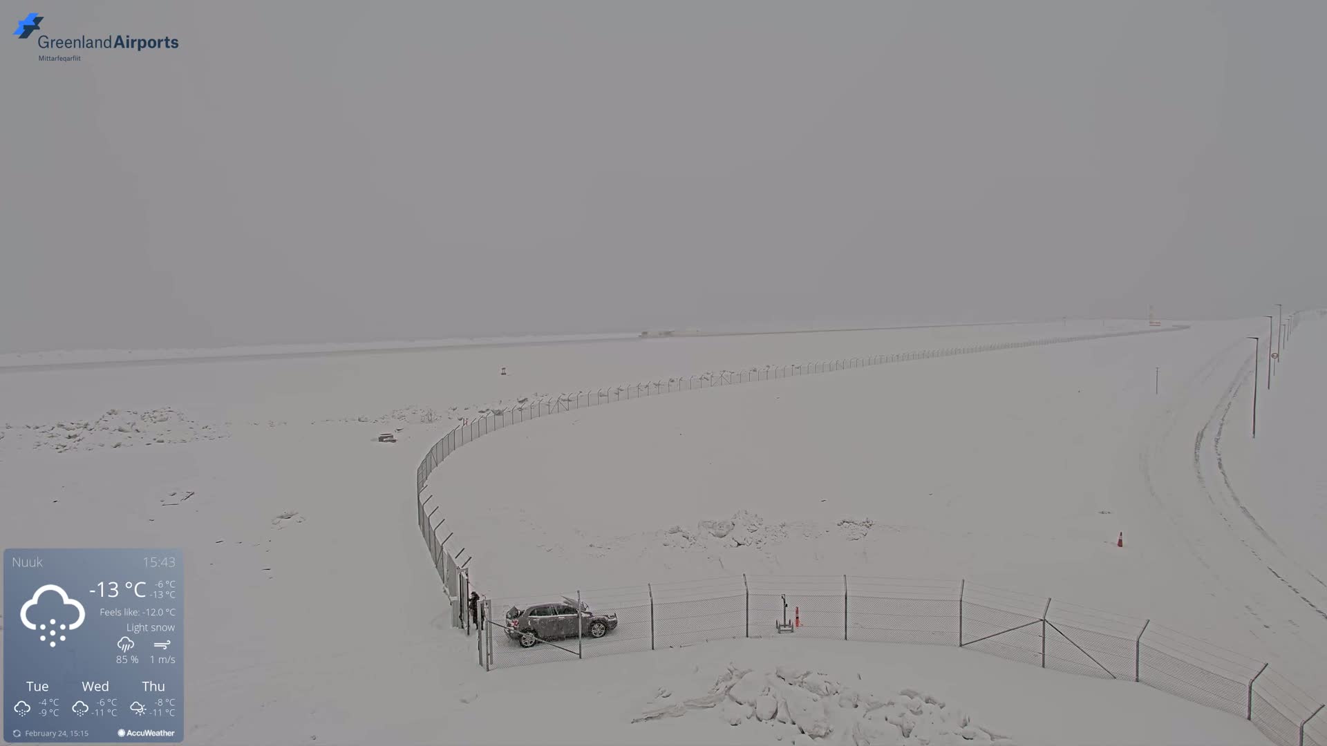 A snow-covered landscape with a chain-link fence, a single car, and a road under light snow in overcast conditions.