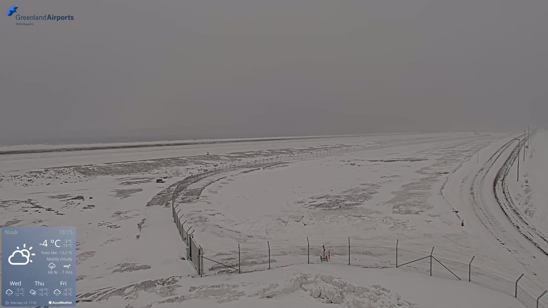 A snow-covered airport runway and surrounding area are visible under mostly cloudy skies.