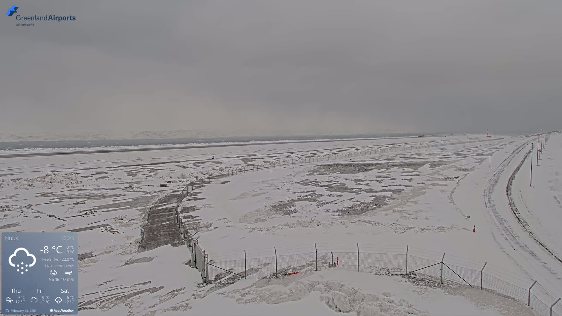 A snow-covered airport runway and surrounding area are visible under a light snow shower during overcast conditions.