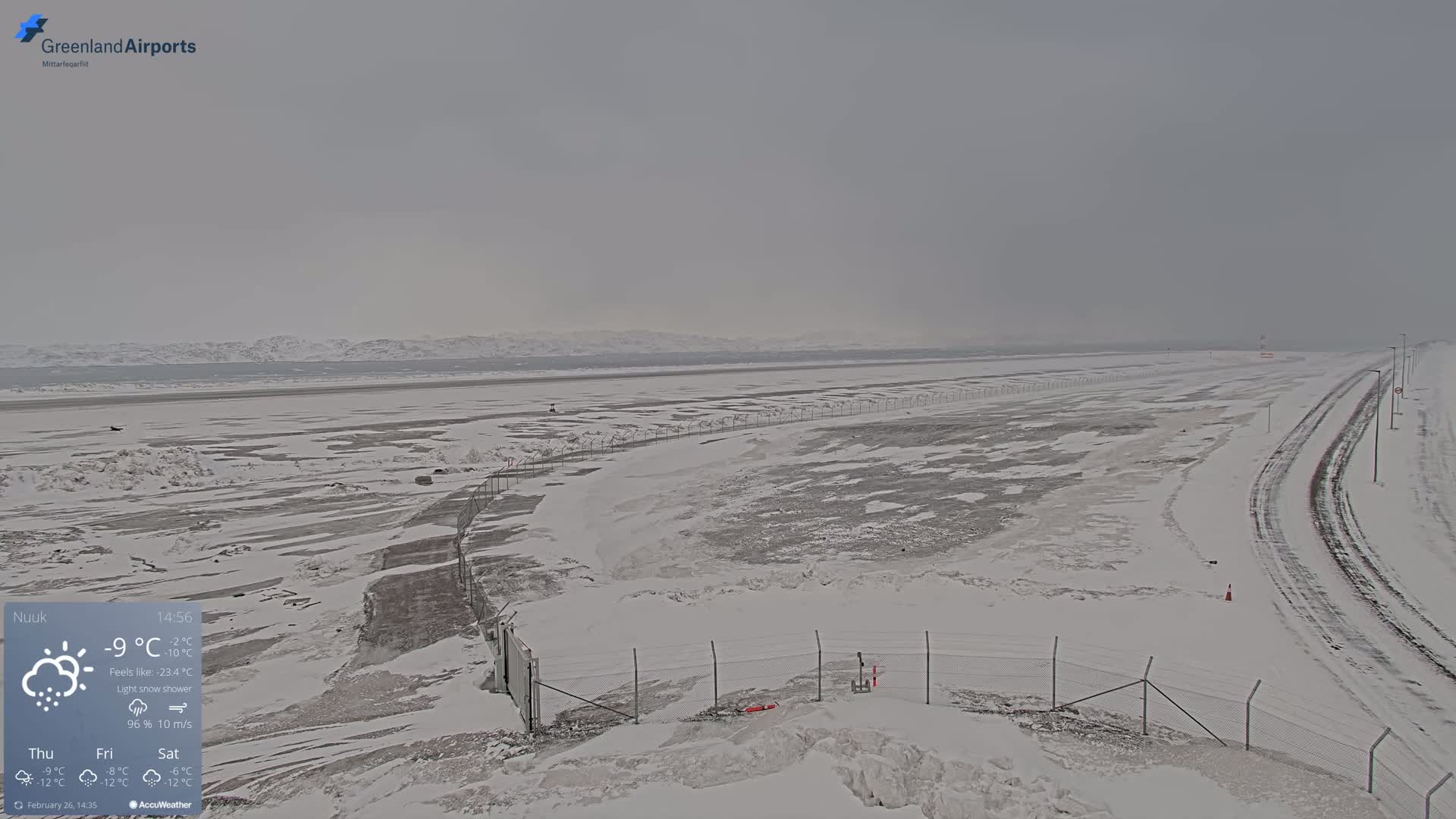 A snow-covered airport runway and surrounding area are visible under a light snow shower with an overcast sky and distant mountains.