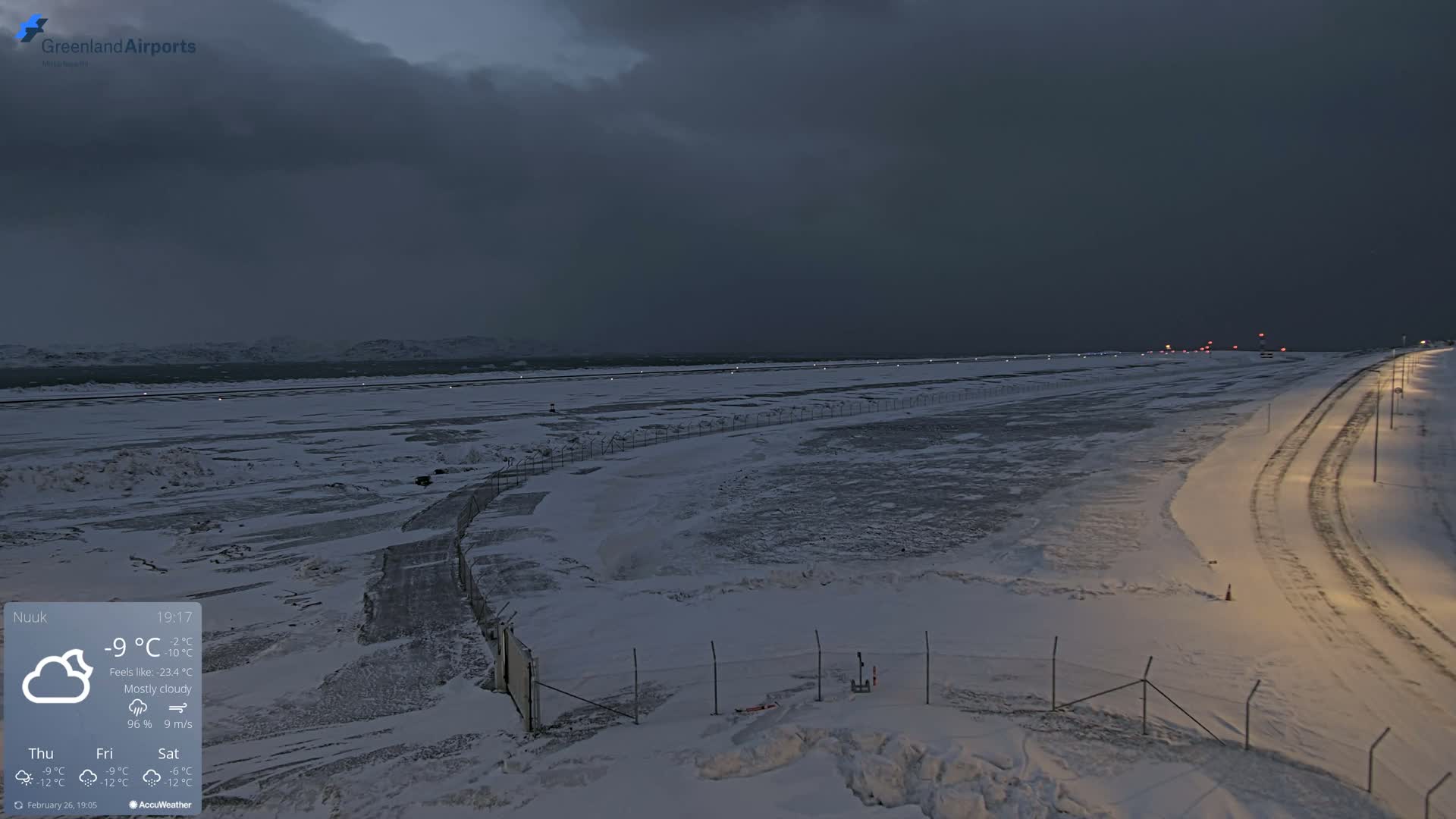 A mostly cloudy, dark evening scene shows a snow-covered airport runway and surrounding area, with a road leading away from the runway.