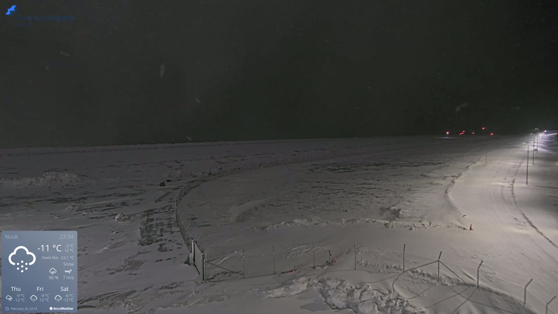 A snow-covered landscape at night is illuminated by lights along a road, with snow falling under a dark sky.