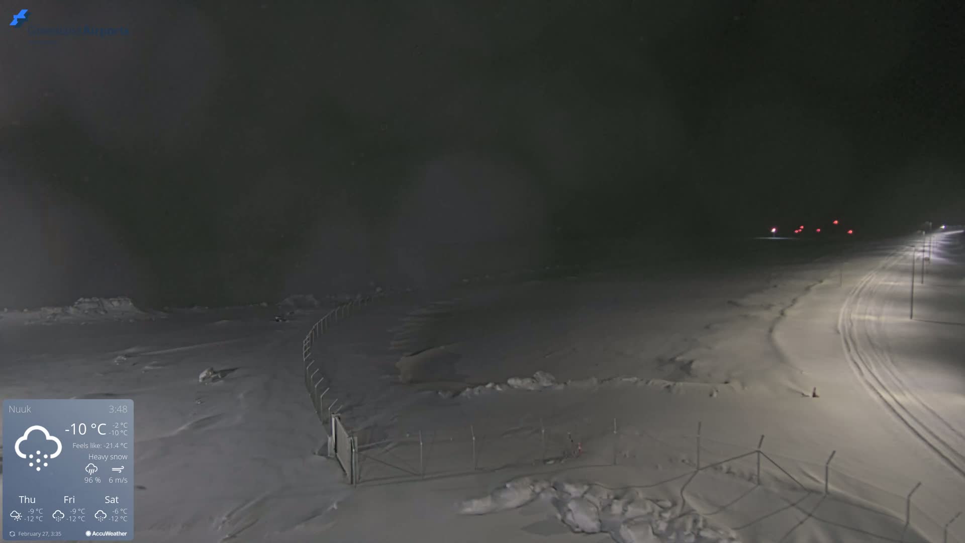 A snow-covered landscape at night is illuminated by distant lights under heavy snowfall.