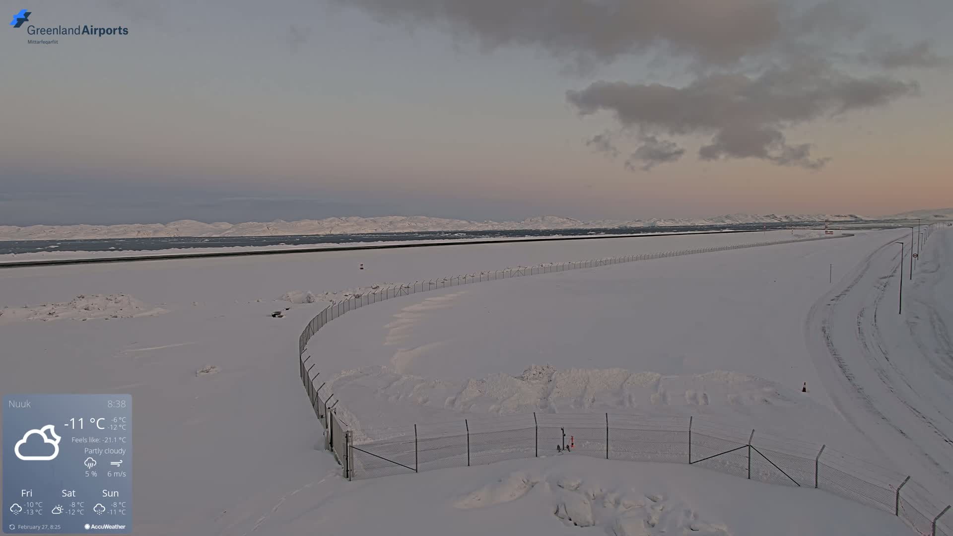 A snow-covered landscape with a chain-link fence and distant mountains under a partly cloudy sky.