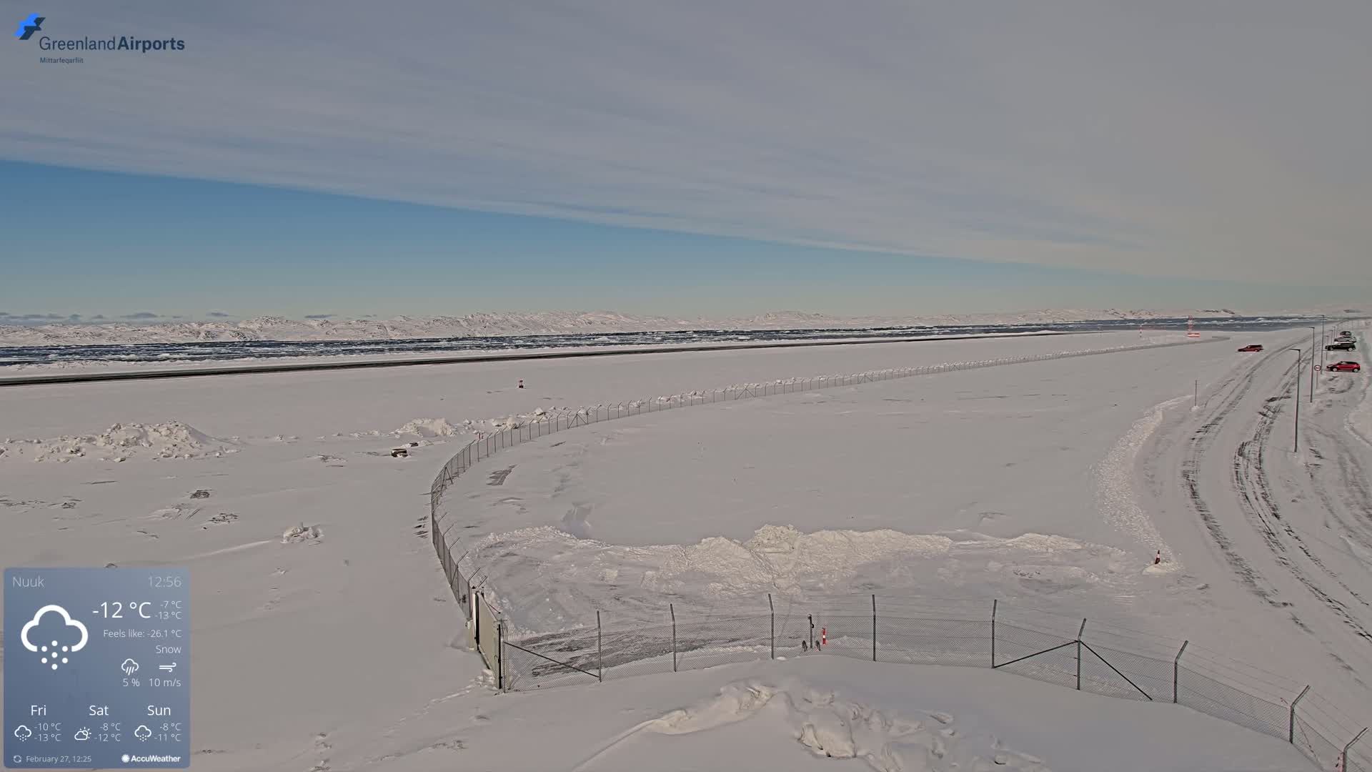 A snow-covered airport runway and surrounding area are visible under a mostly clear, bright sky with a few wispy clouds.