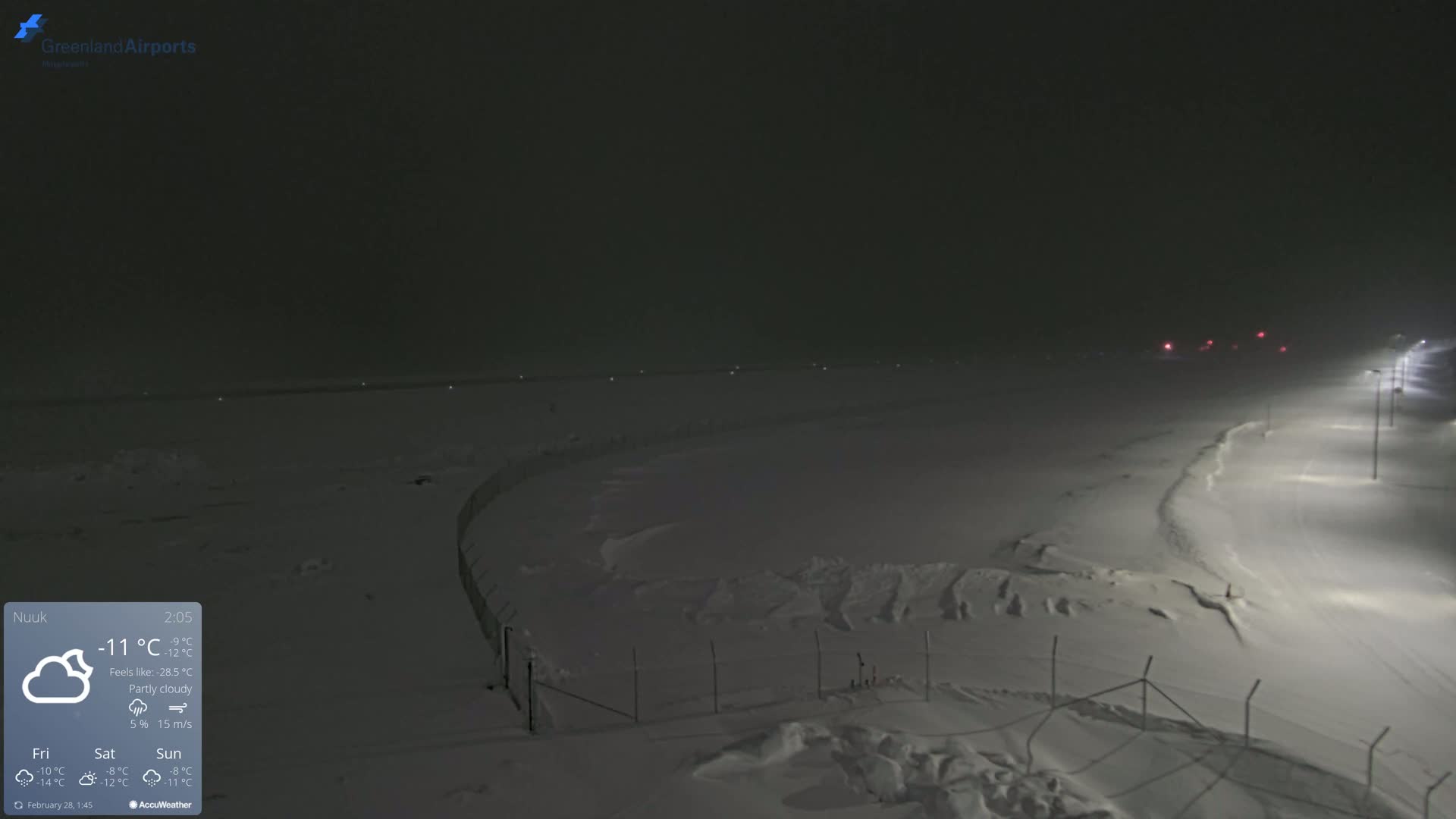 A snow-covered, partly cloudy nighttime scene shows a fenced-in area illuminated by lights, with a distant runway faintly visible.