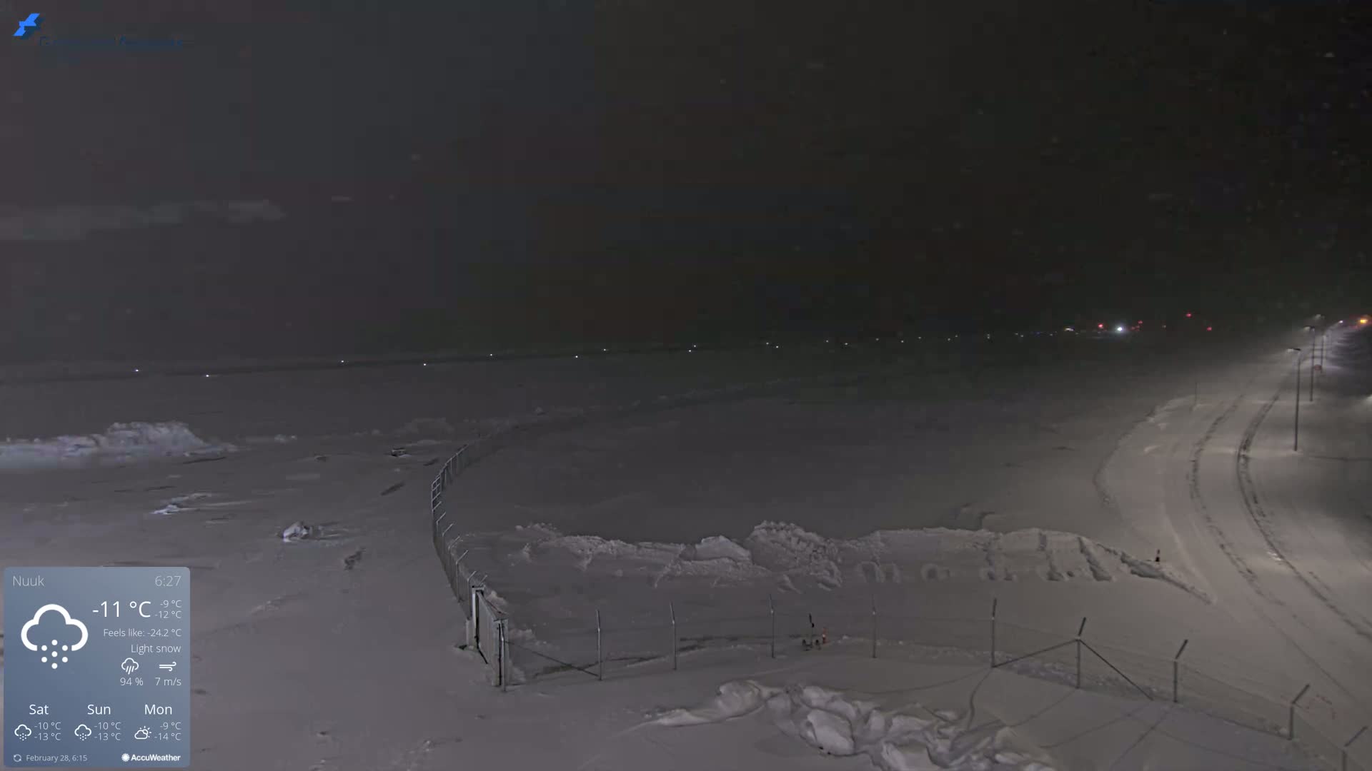 A snow-covered landscape at night with light snow falling, illuminated by streetlights along a road, and a fence separating the road from a snow-covered field.