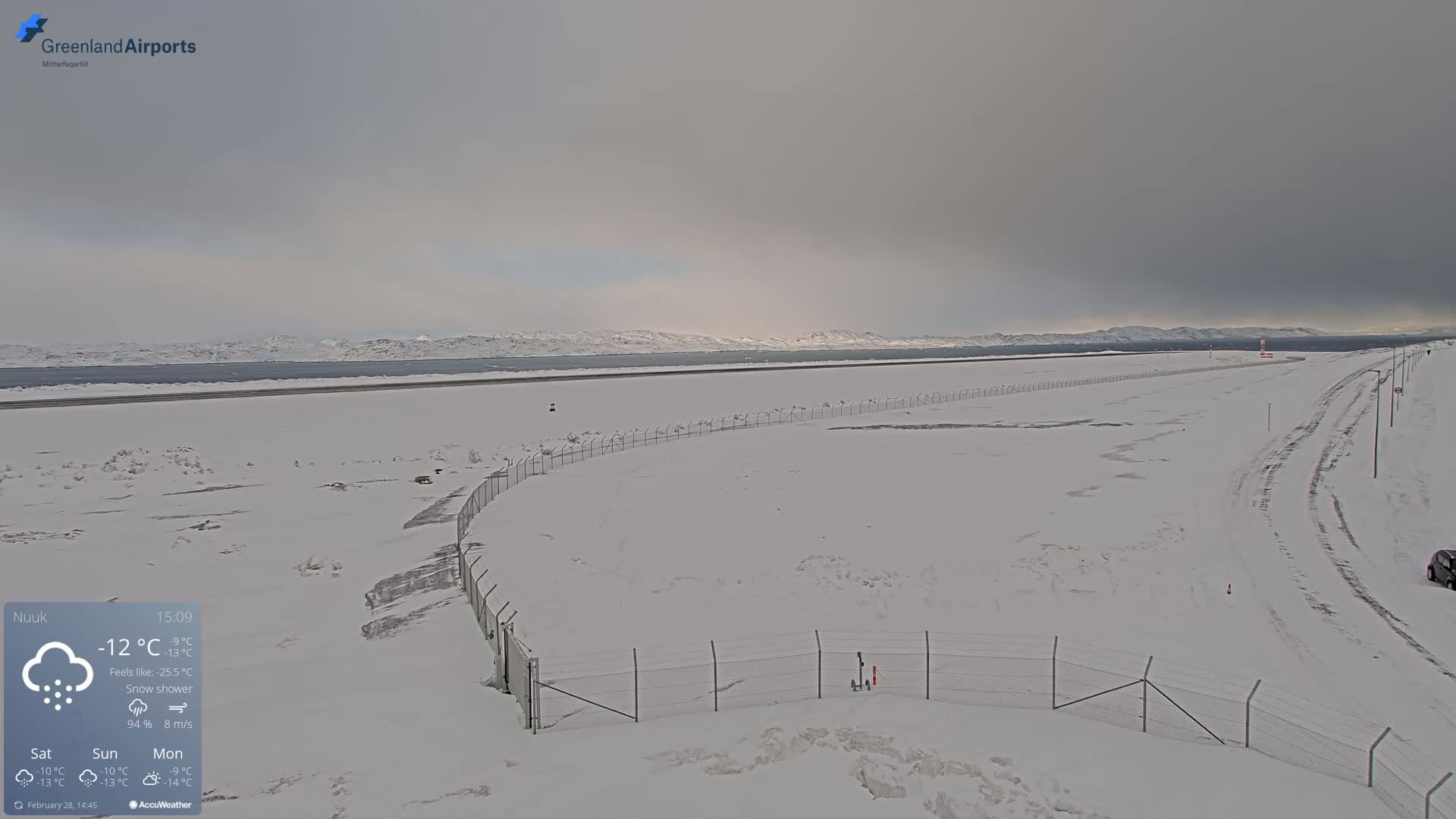 A snow-covered airport runway and surrounding area are visible under a cloudy sky, with mountains in the distance.