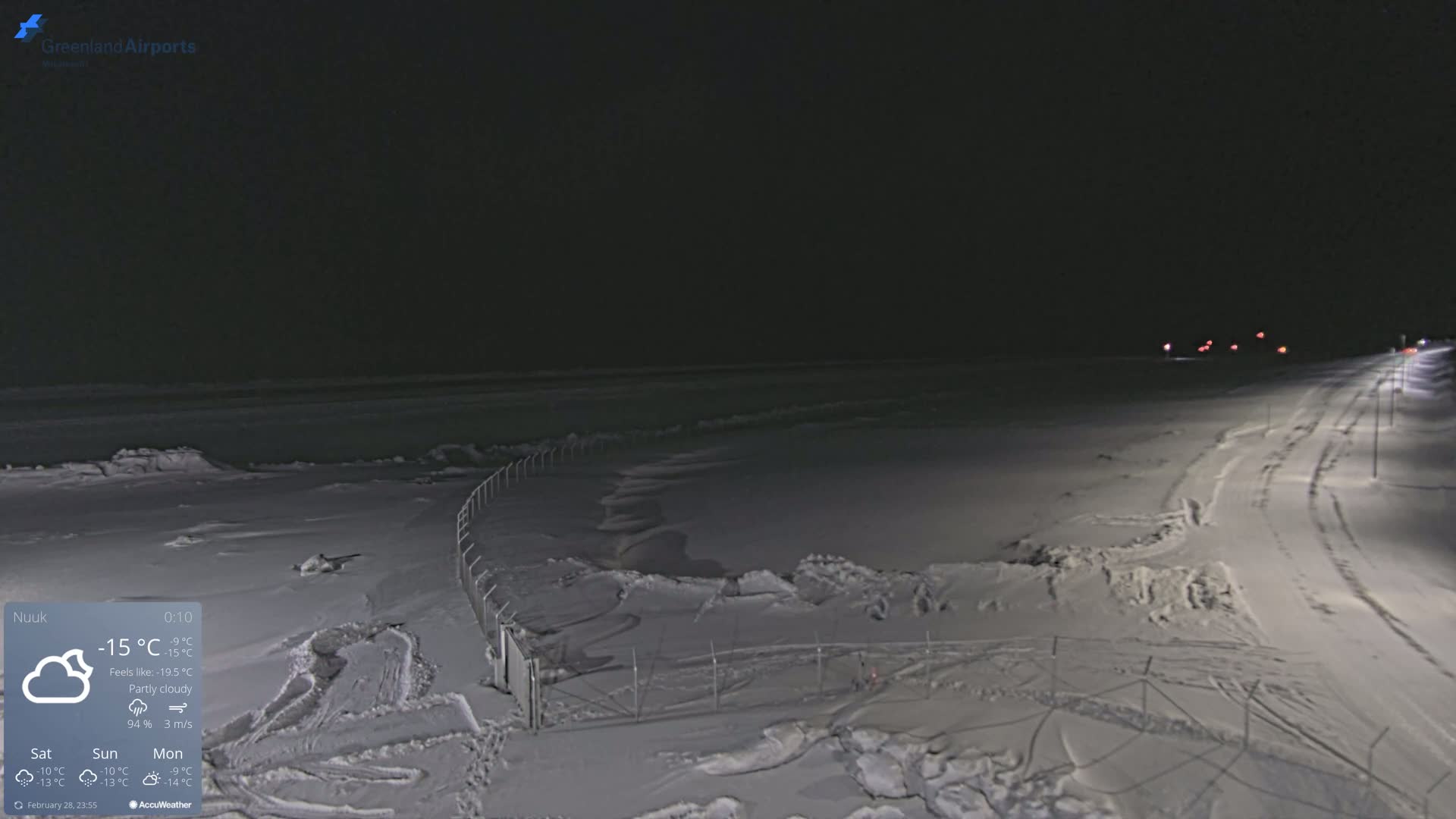 A snow-covered landscape at night, illuminated by lights along a road, with a fence in the foreground under partly cloudy skies.