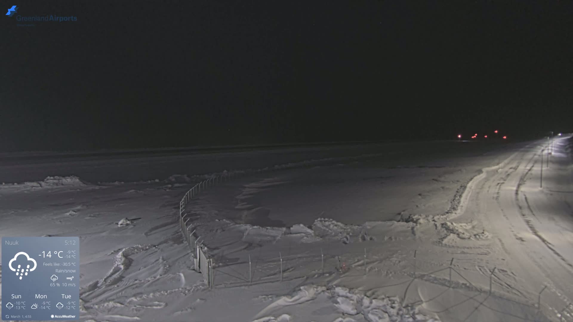 A snow-covered, dark landscape at night shows a fenced area, a road with faint tire tracks, and distant lights under a rain/snow mix.