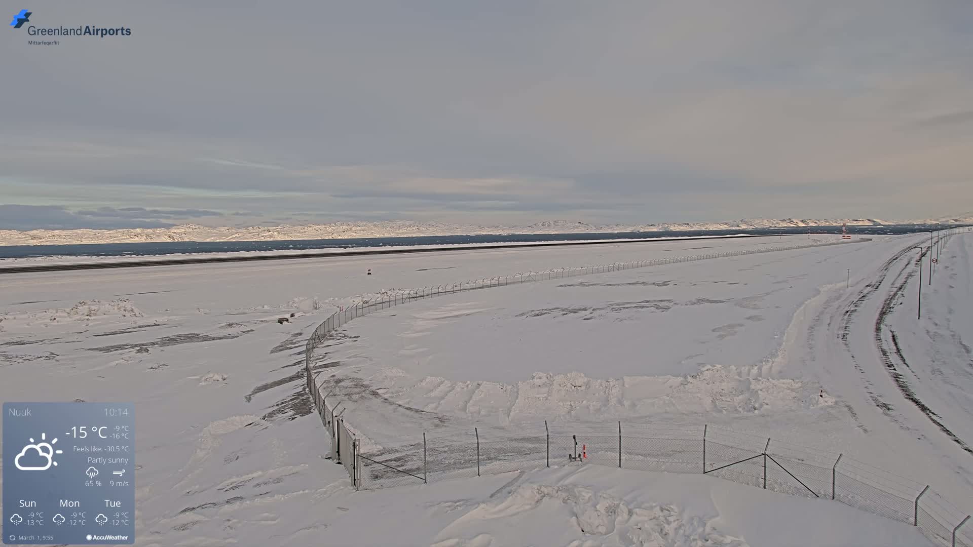 A snow-covered airport runway stretches towards a distant, snow-capped mountain range under a partly sunny sky.