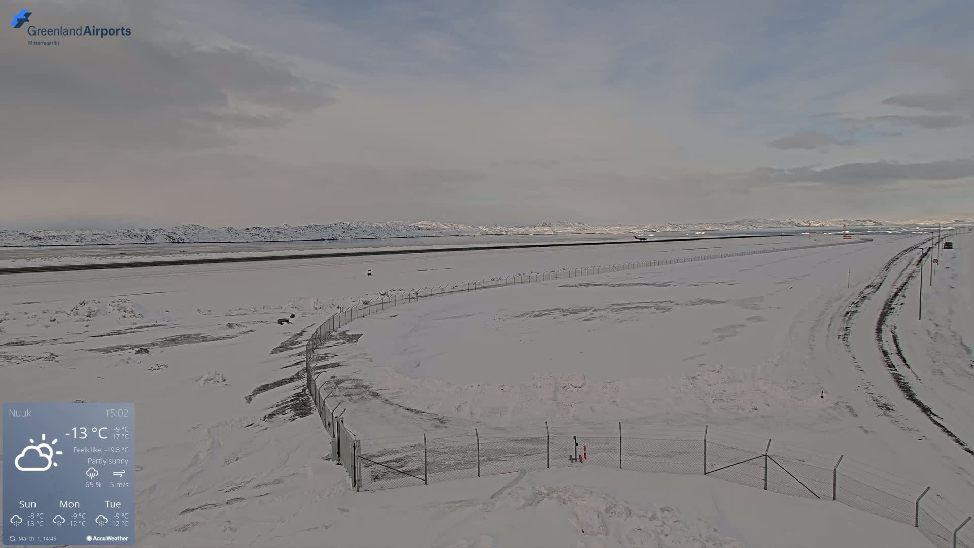 A snow-covered airport tarmac and runway extend to a partly sunny sky and distant, snow-capped mountains.