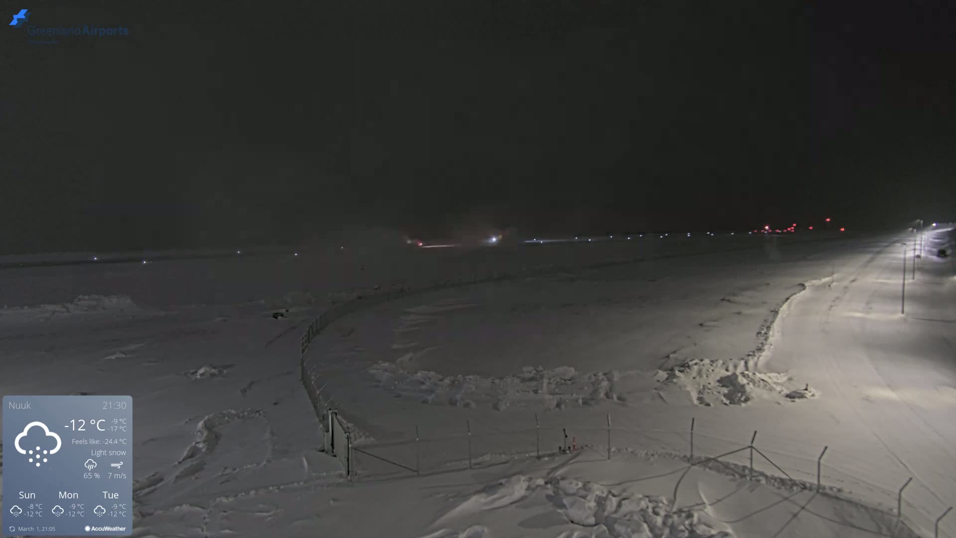 A snow-covered airport runway and surrounding area are dimly lit at night during a light snow.
