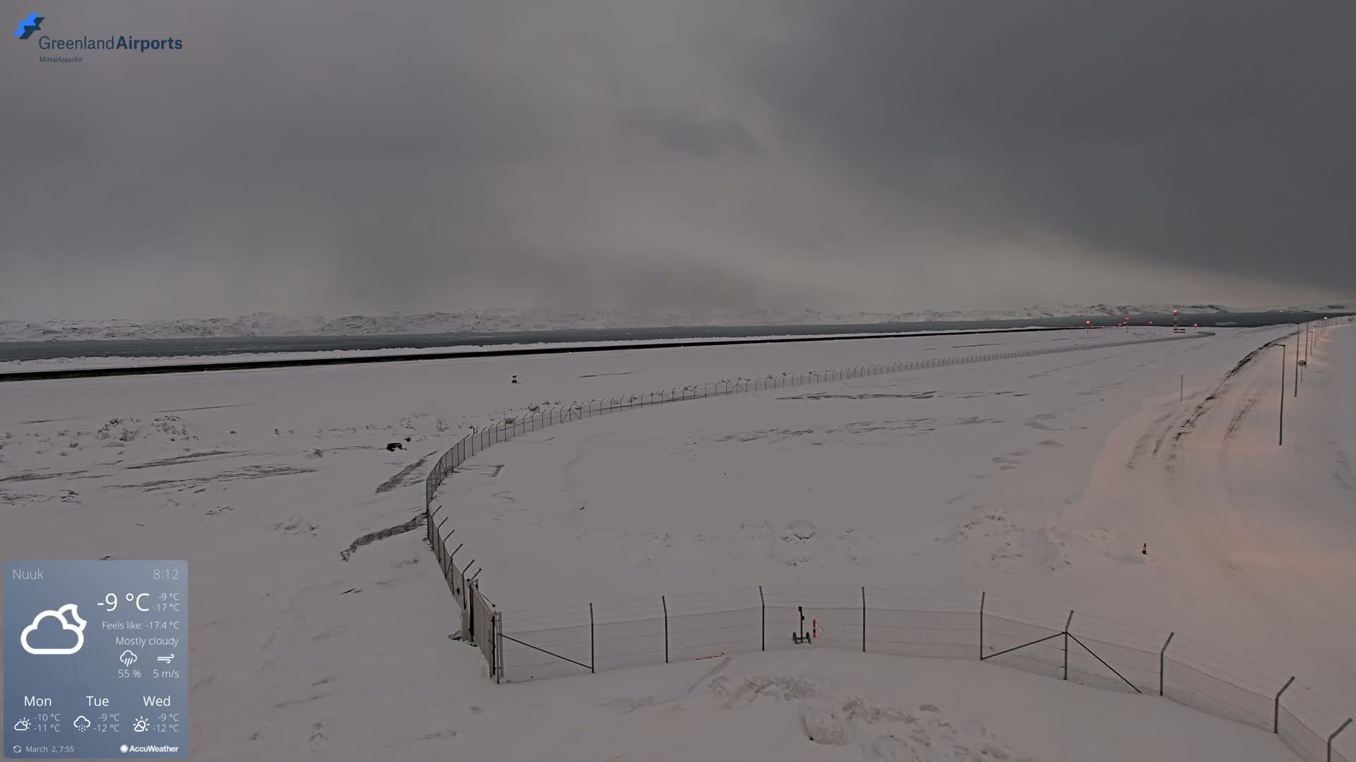 A snow-covered airport runway extends to a mostly cloudy, gray sky and distant mountains.