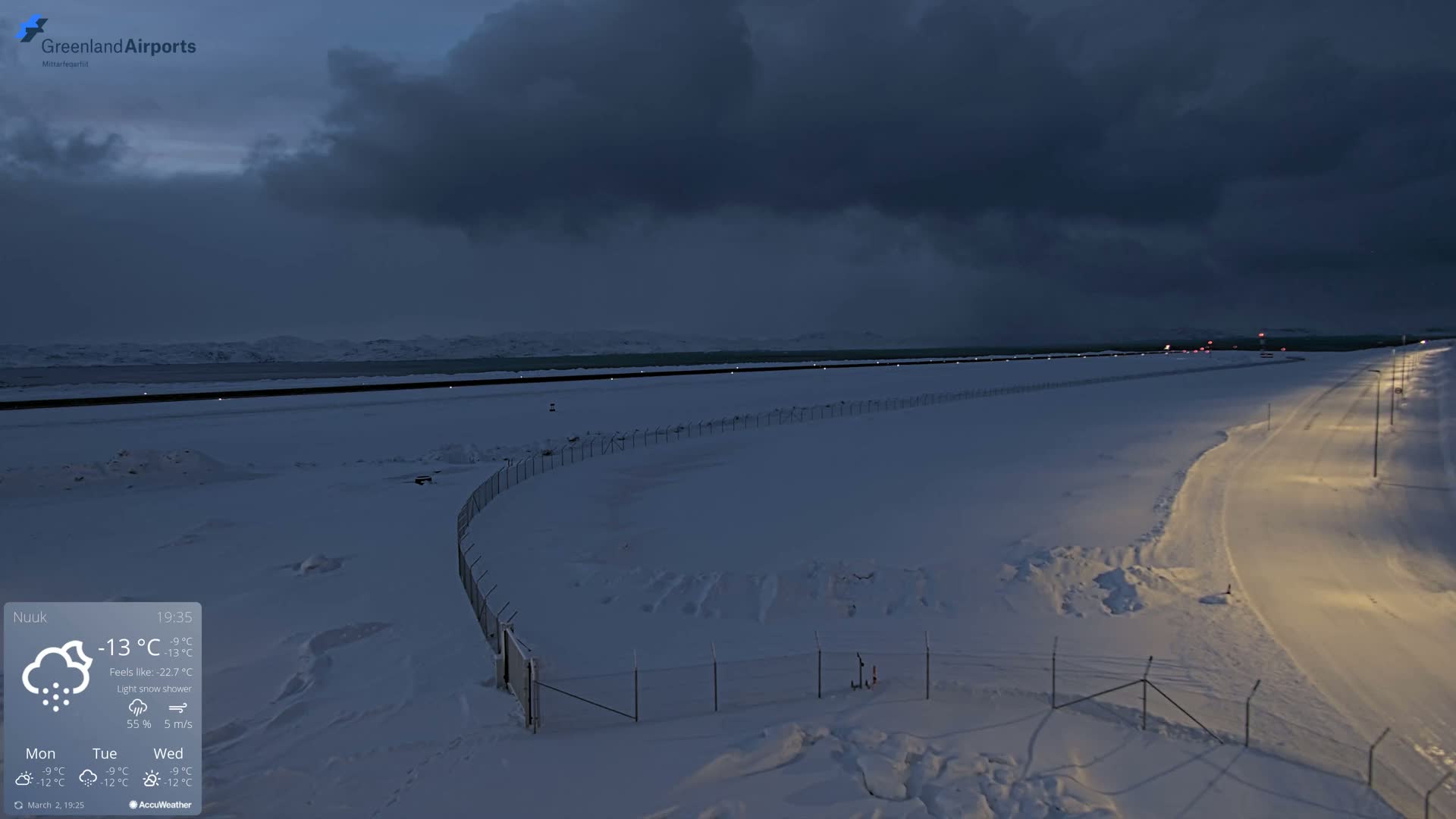 A snow-covered airport runway and road are visible under a dark, cloudy sky during a light snow shower.