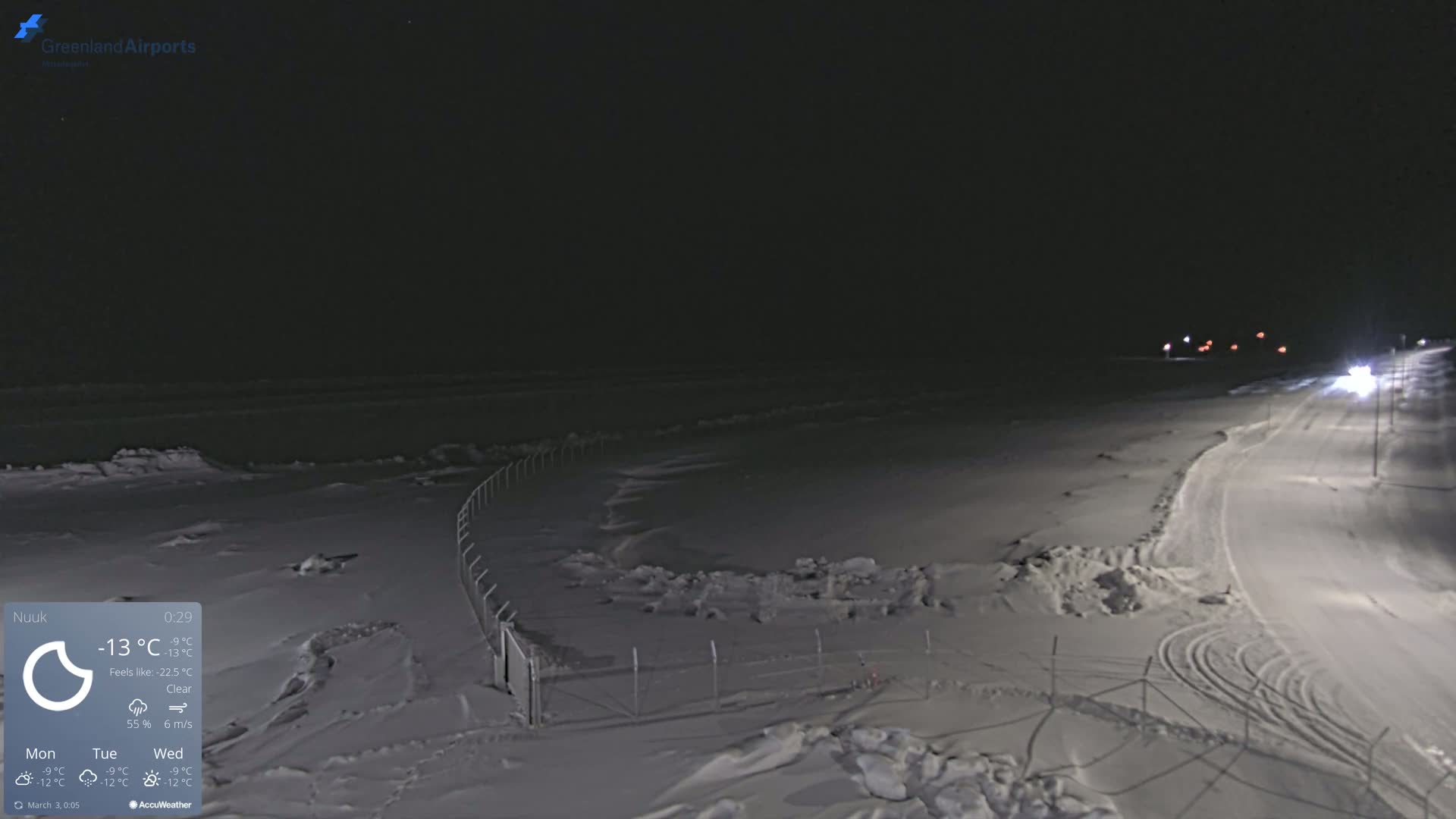 A snow-covered landscape at night, illuminated by distant lights, with a clear sky and a fence along a road.