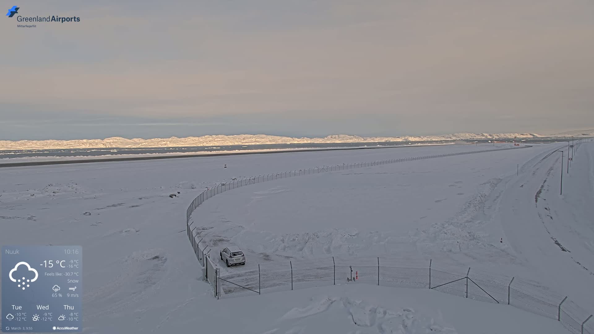 A snow-covered airfield with a single car parked near a fence, mountains in the distance under a pale sky with light snowfall.