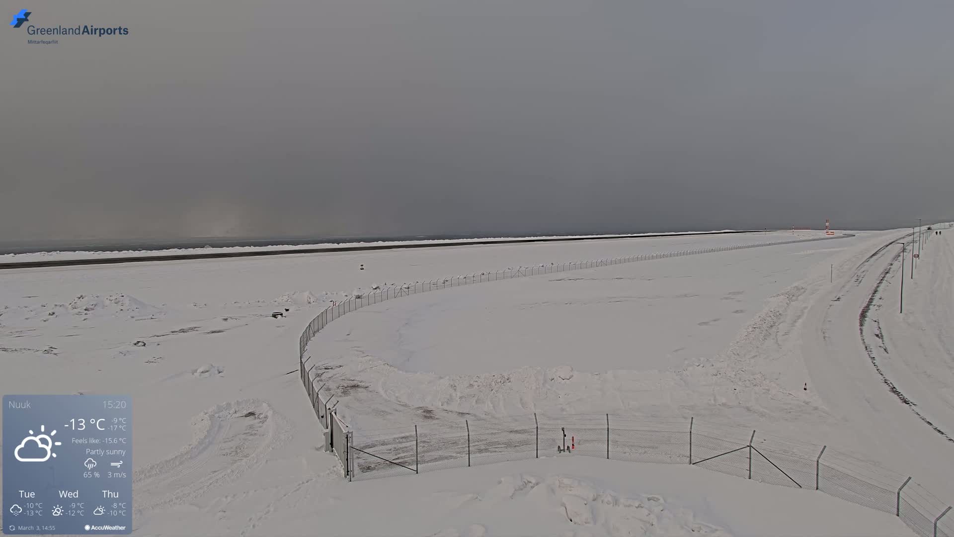 A snow-covered airport runway and surrounding area are visible under a partly sunny, overcast sky.