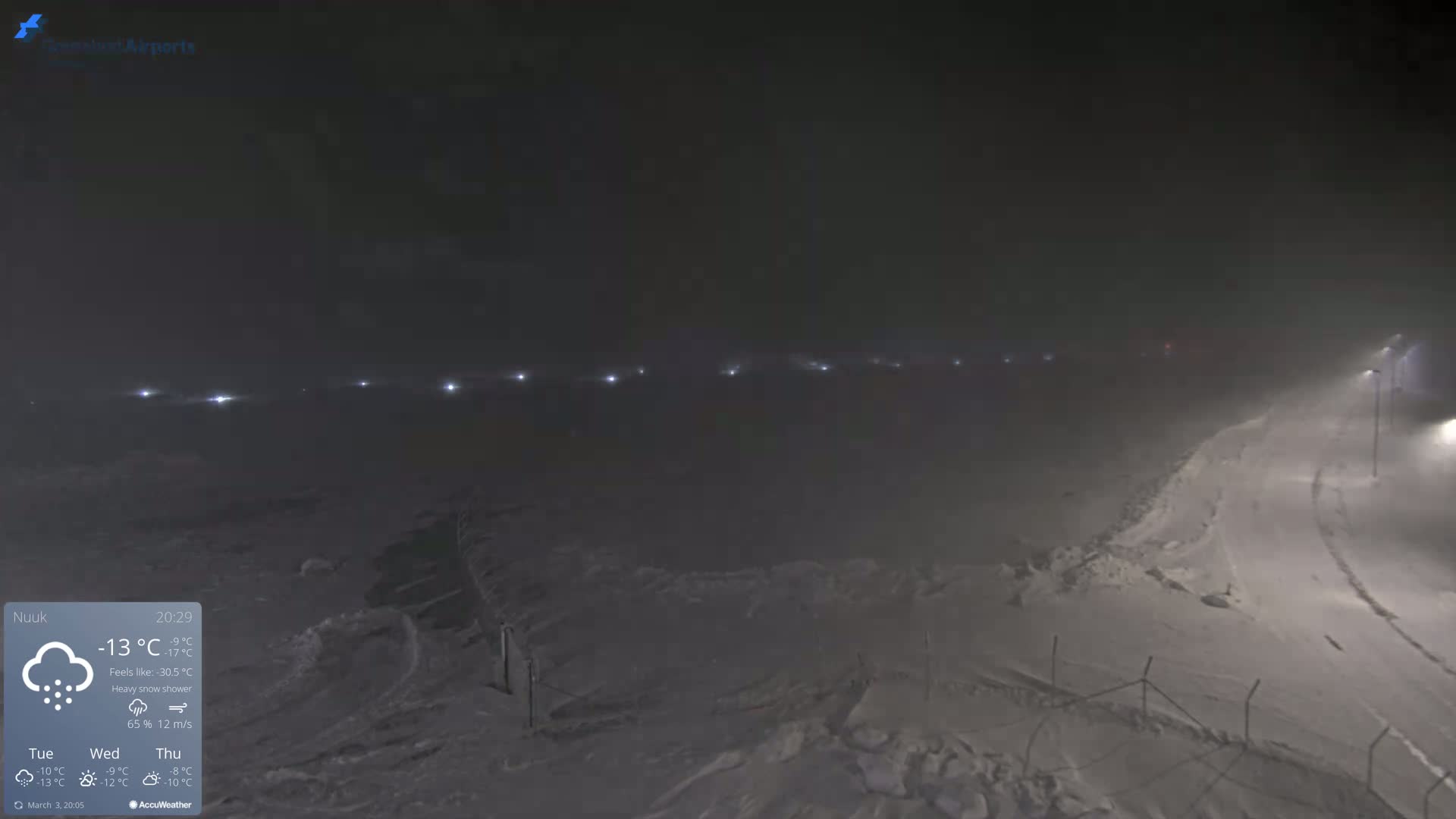 A snow-covered landscape at night is illuminated by a line of distant lights in heavy snow and wind.