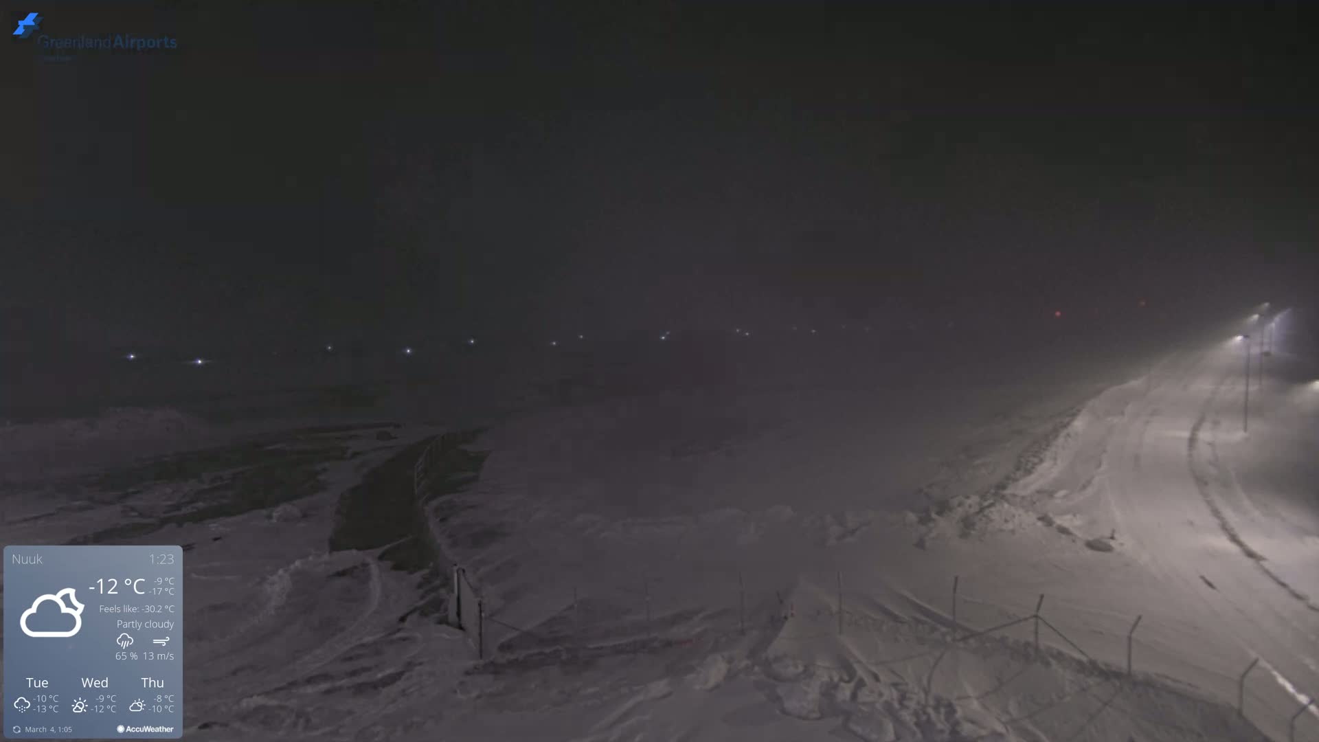 A snow-covered landscape at night is dimly lit by several lights in the distance under a partly cloudy sky.