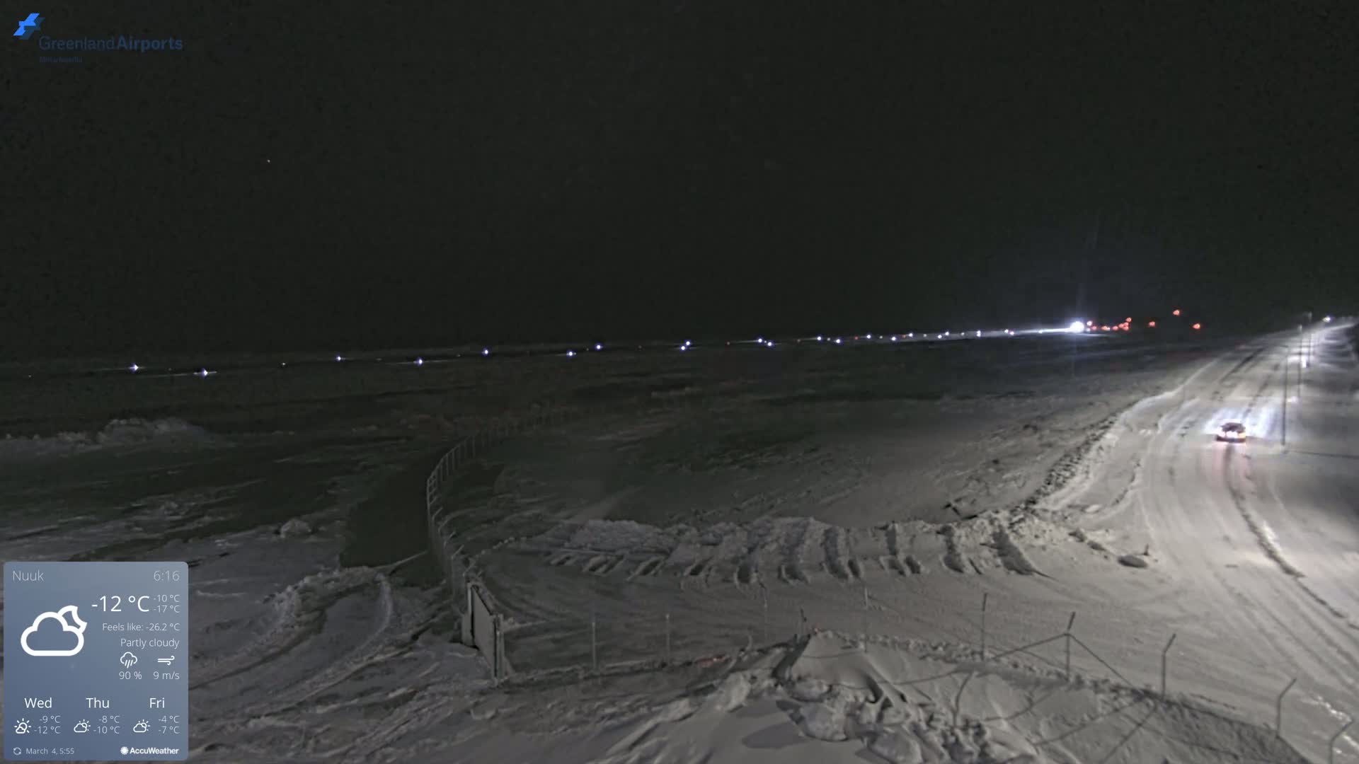 A snow-covered airfield at night under partly cloudy skies, with a vehicle driving on a nearby road.