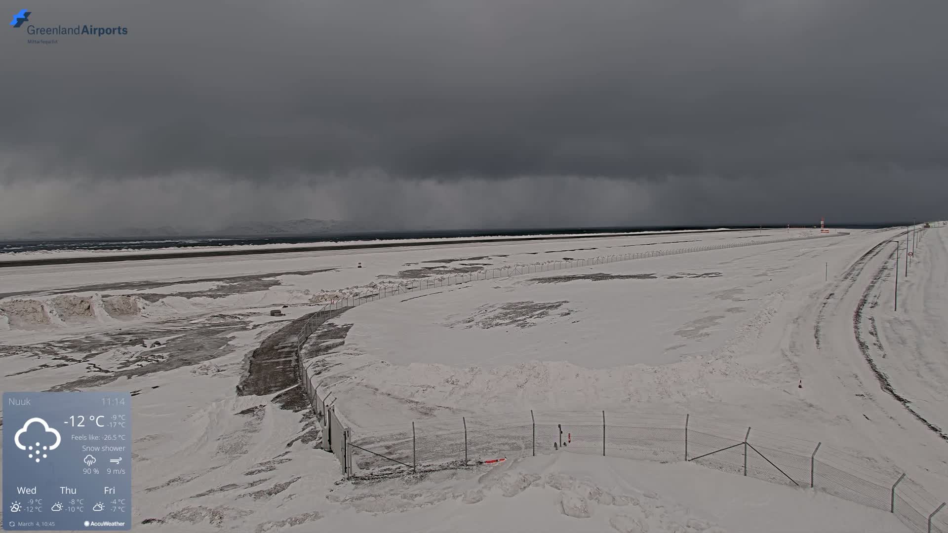 A snow-covered airport runway and surrounding area are visible under a dark, overcast sky with a snow shower.