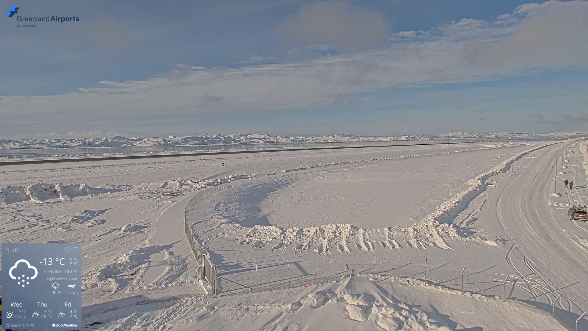 A snow-covered airport runway and surrounding area extend to a frozen body of water and distant mountains under a partly cloudy sky, with light snow falling.