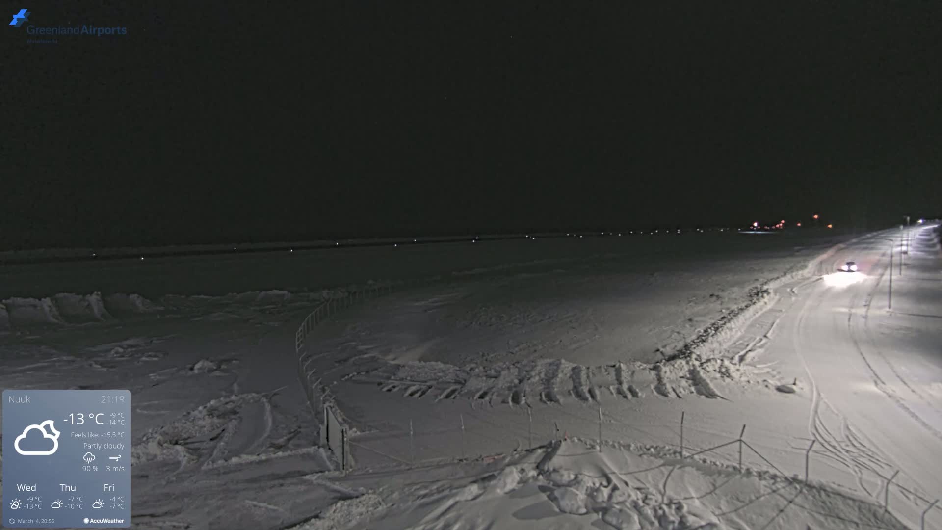 A snow-covered landscape at night, under a partly cloudy sky, with a vehicle driving on a lit road.