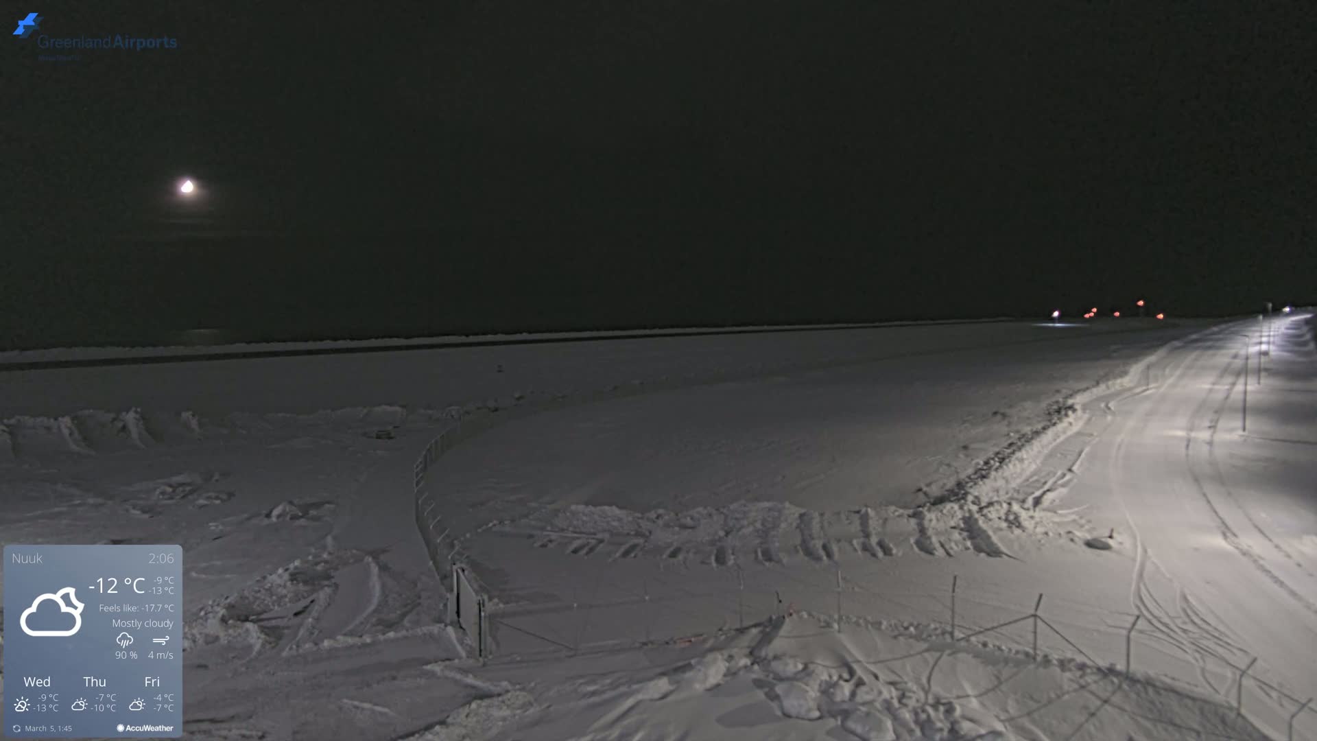 A mostly cloudy night scene shows a snow-covered landscape with a road and fence under a bright moon.