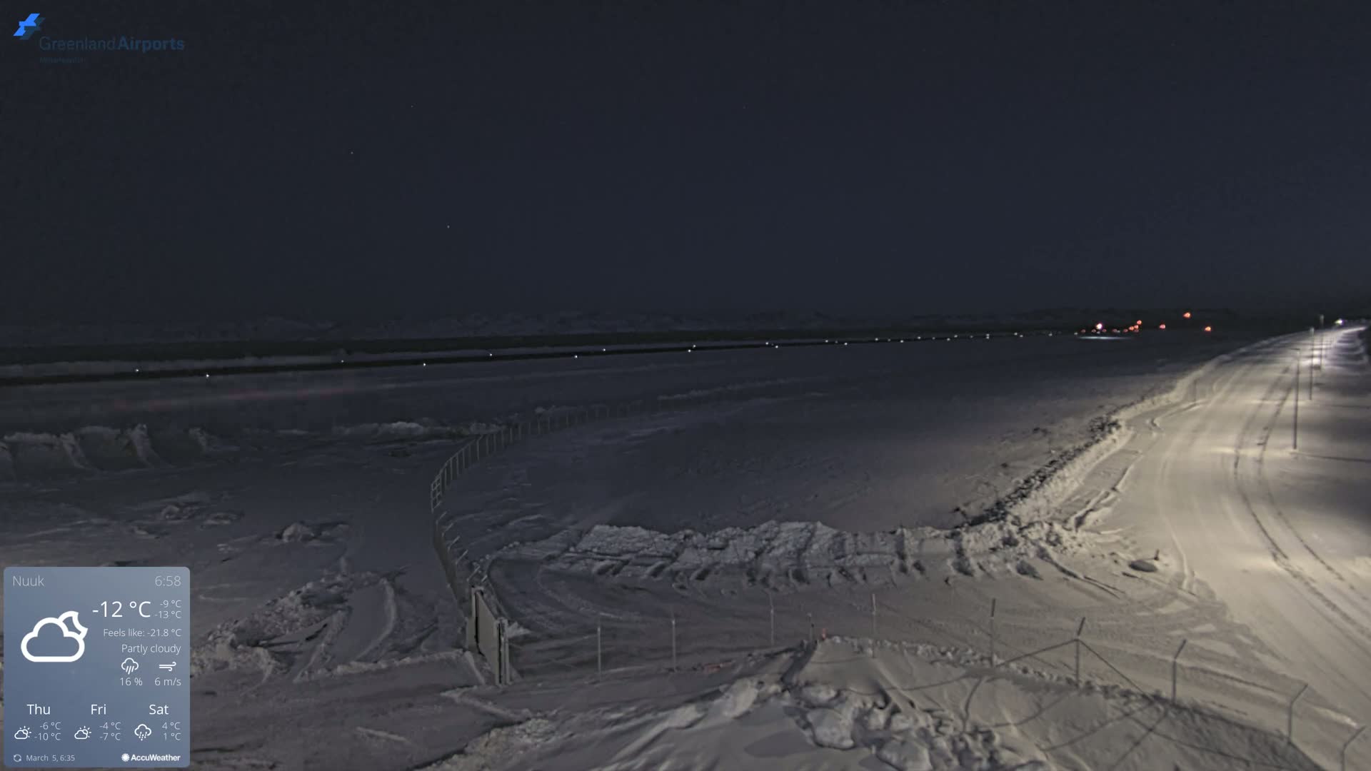 A snow-covered landscape at night, partially illuminated by lights along a road and a fence, under partly cloudy skies.