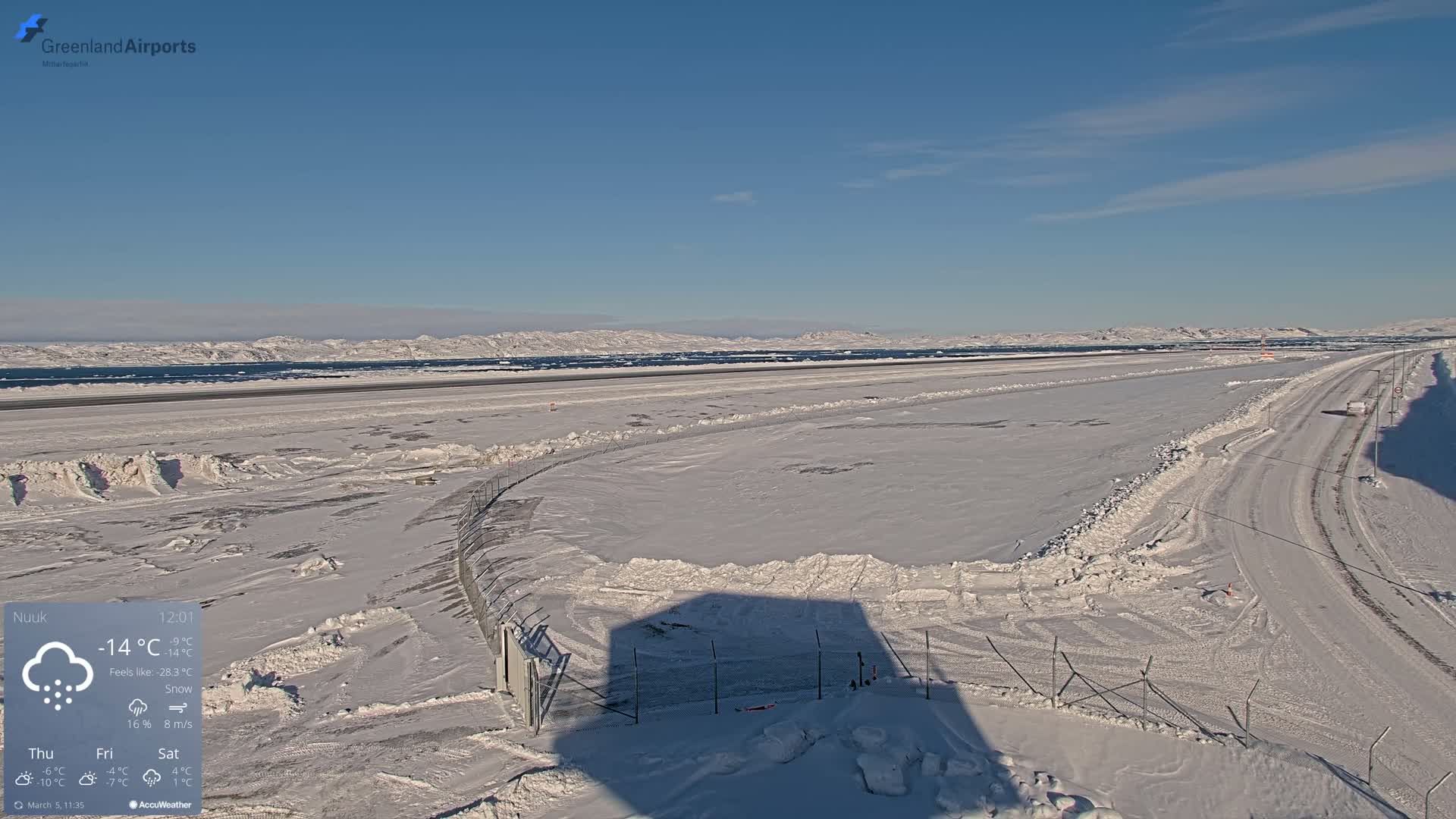 A snow-covered airport runway and road extend toward a distant, snow-covered mountain range under a clear, blue sky.