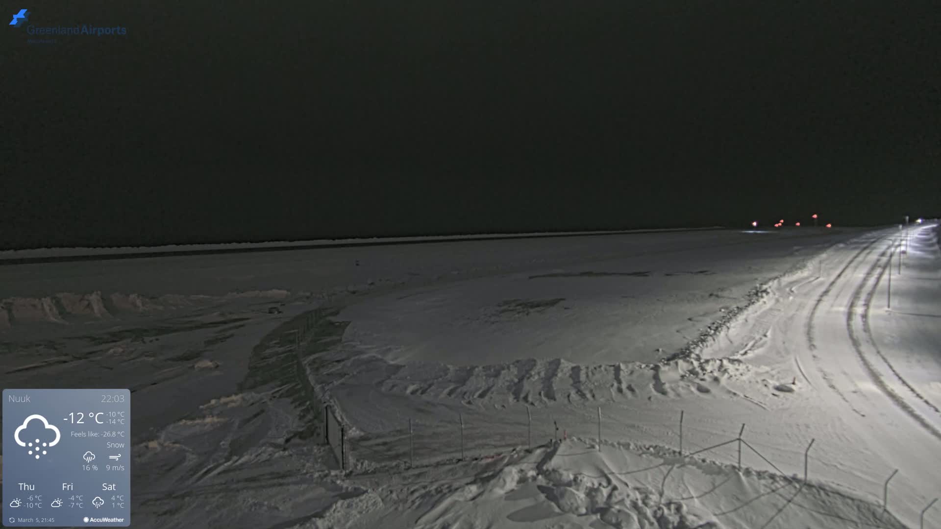 A snow-covered, flat landscape at night, with a road and faint lights visible in the distance under a dark sky.