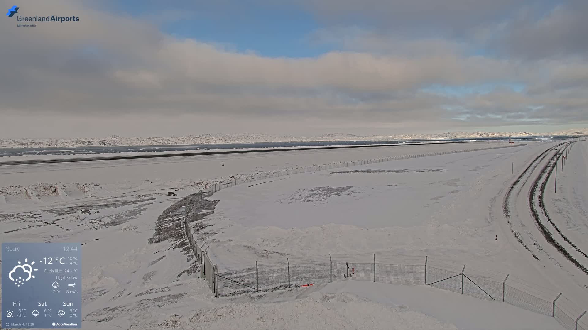 A snow-covered airport runway and surrounding area are visible under a cloudy sky with light snow.