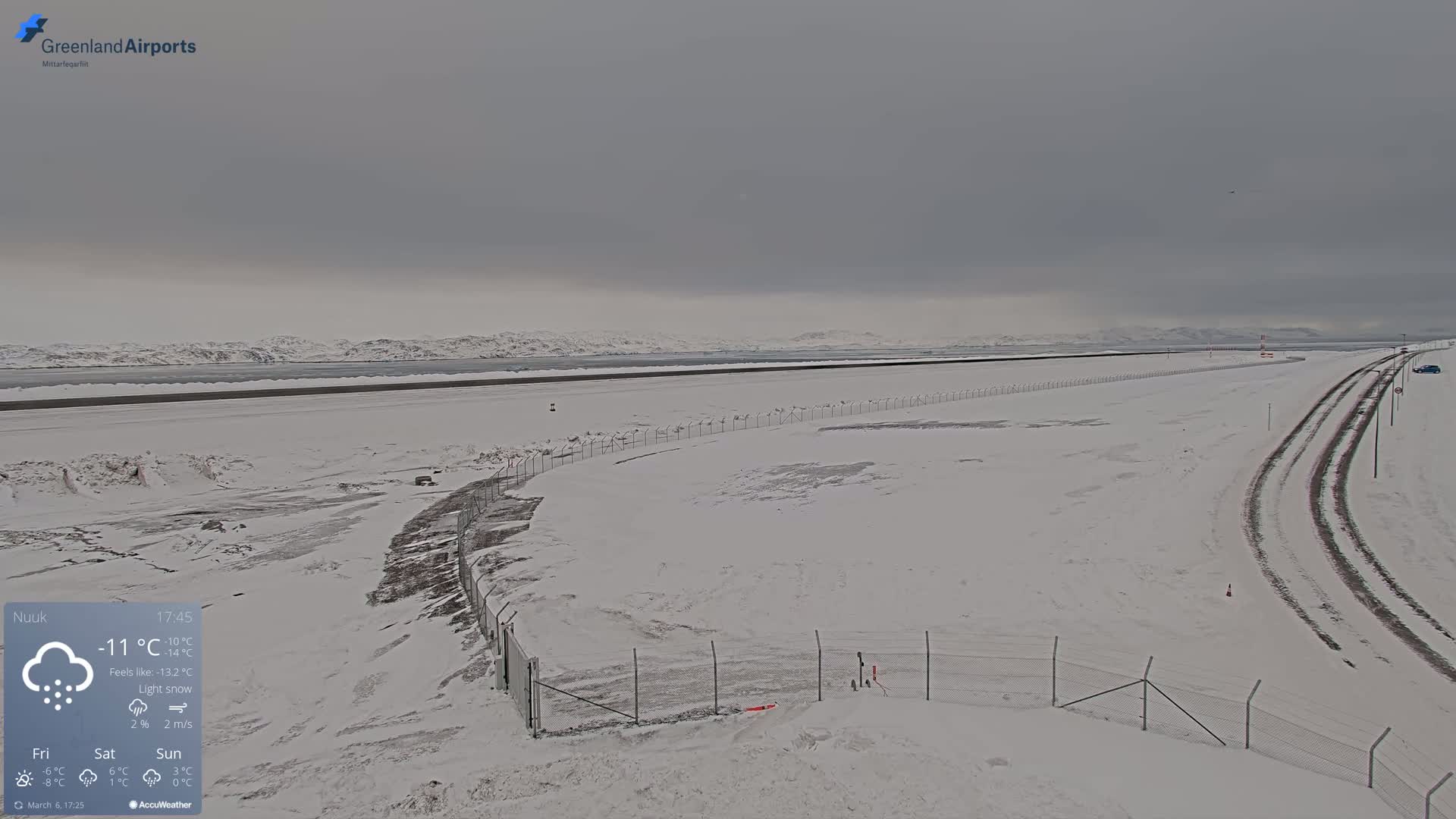 A snow-covered airport runway and surrounding area are visible under a gray, overcast sky with light snow.