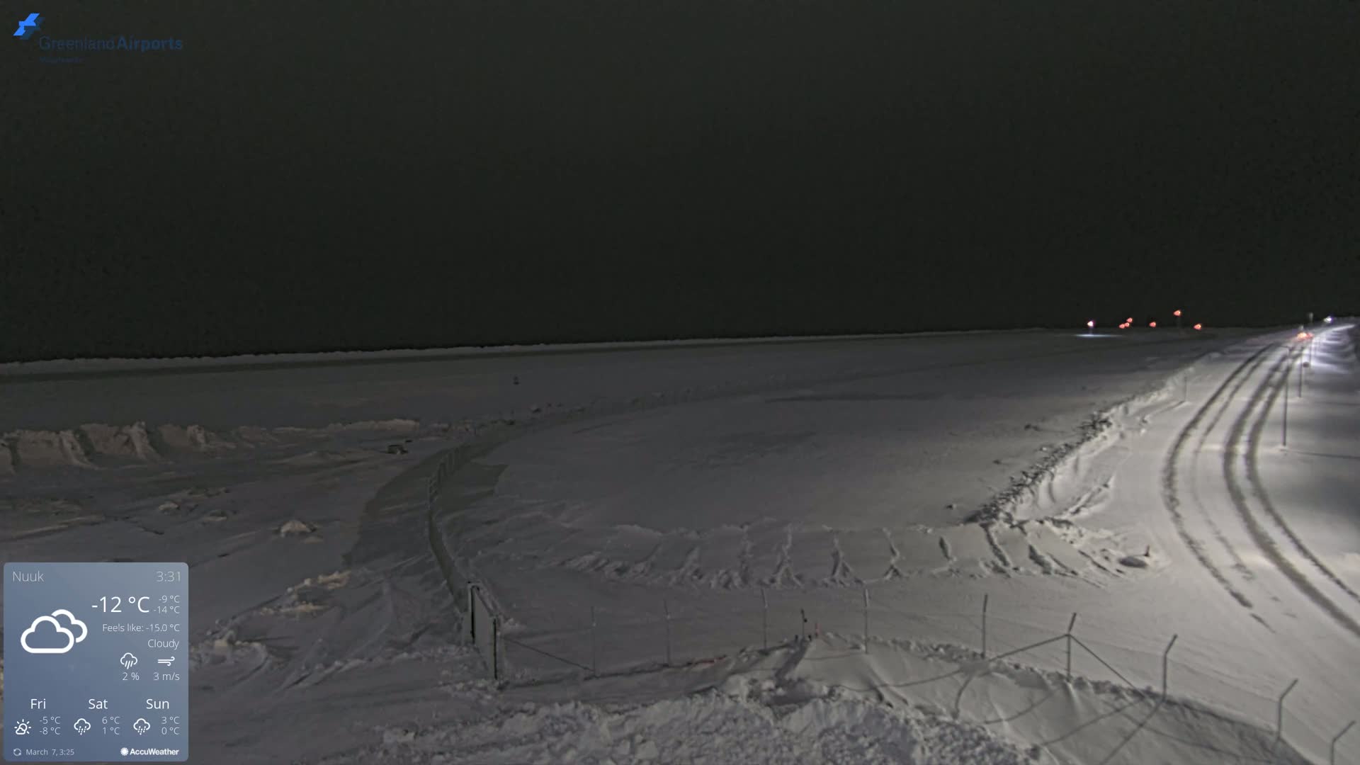 A snow-covered landscape at night, under cloudy skies, shows a road with tire tracks leading toward some distant lights.