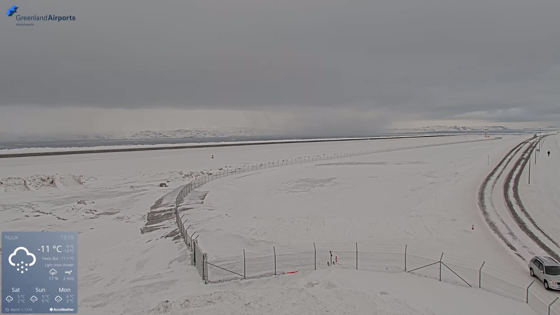 A snow-covered landscape with a road and fence, under a light snow shower and overcast sky, shows a body of water and distant mountains in the background.