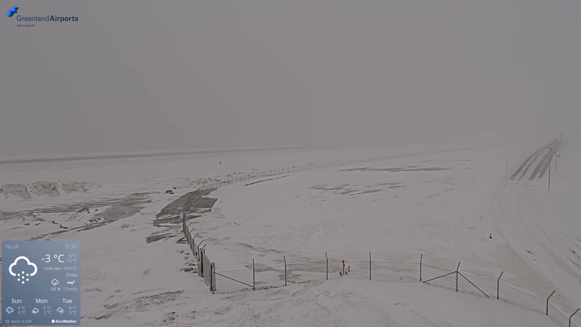 A snow-covered landscape with a chain-link fence and a road visible in the distance under heavy snow.