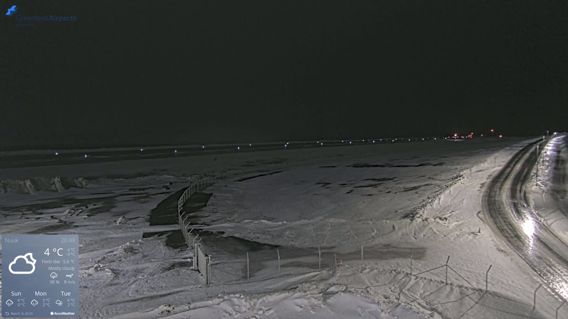 A snow-covered, mostly cloudy nighttime scene shows a road curving through a fenced-in area near a body of water with faint lights along the horizon.