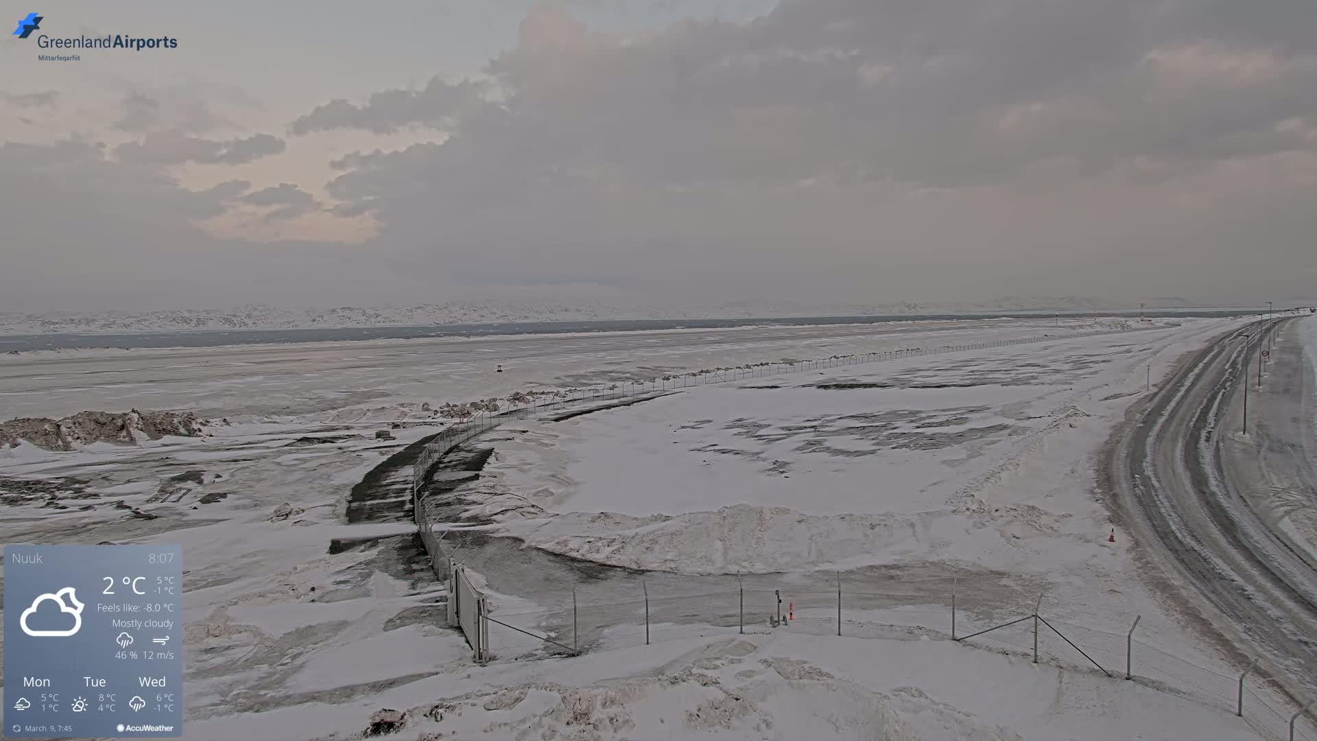 A mostly cloudy, snowy landscape shows a road and fence curving toward a body of water with distant snow-covered mountains under a pale sky.