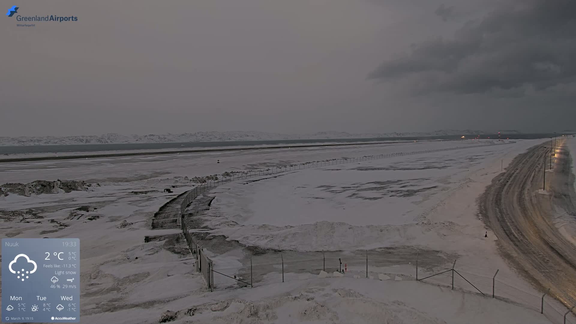 A snow-covered airport runway and road are visible under a cloudy, dark sky with light snow falling.