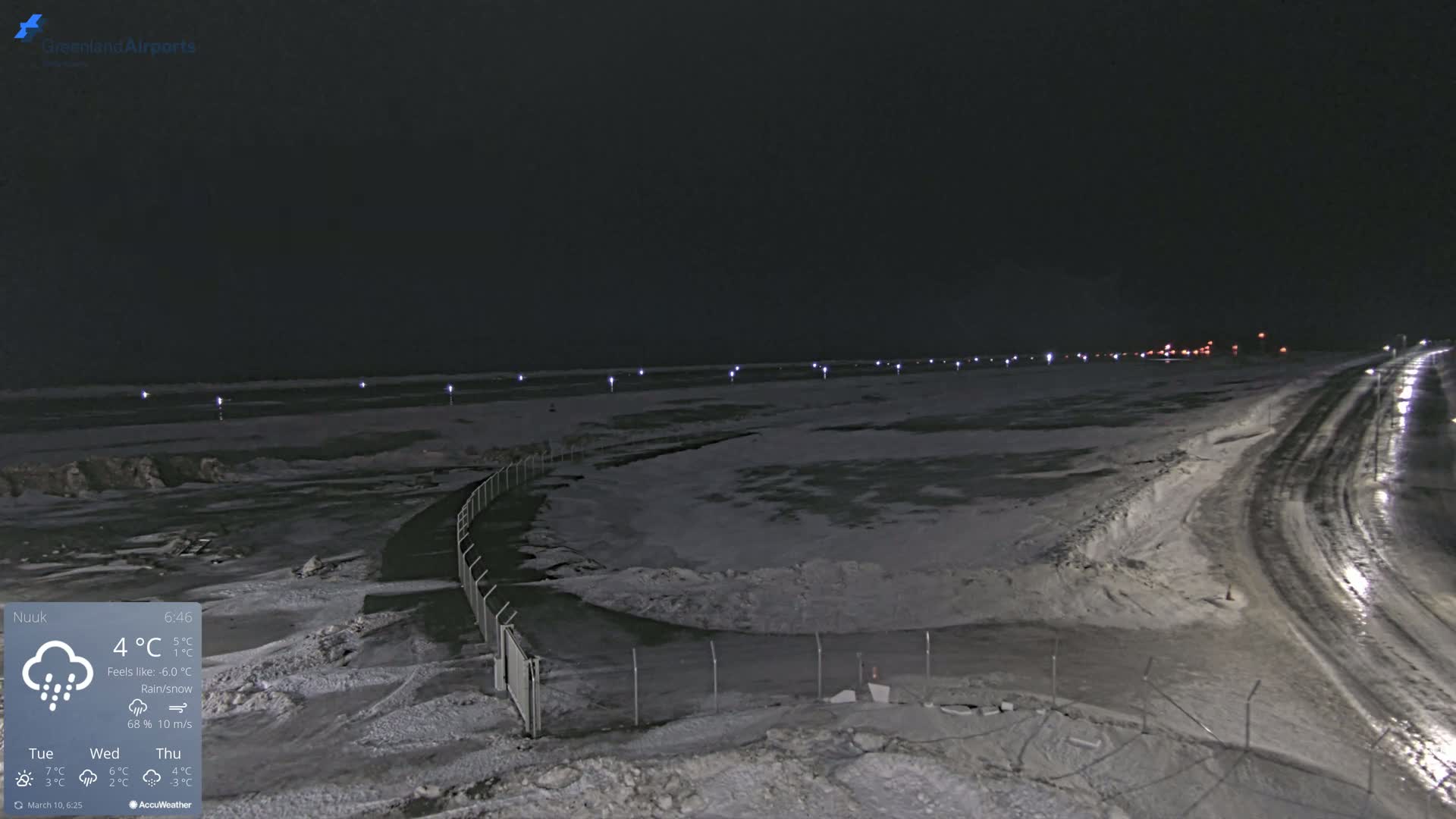 A snow-covered, dark landscape at night with a road and a fence, illuminated by lights, under conditions of rain and snow.