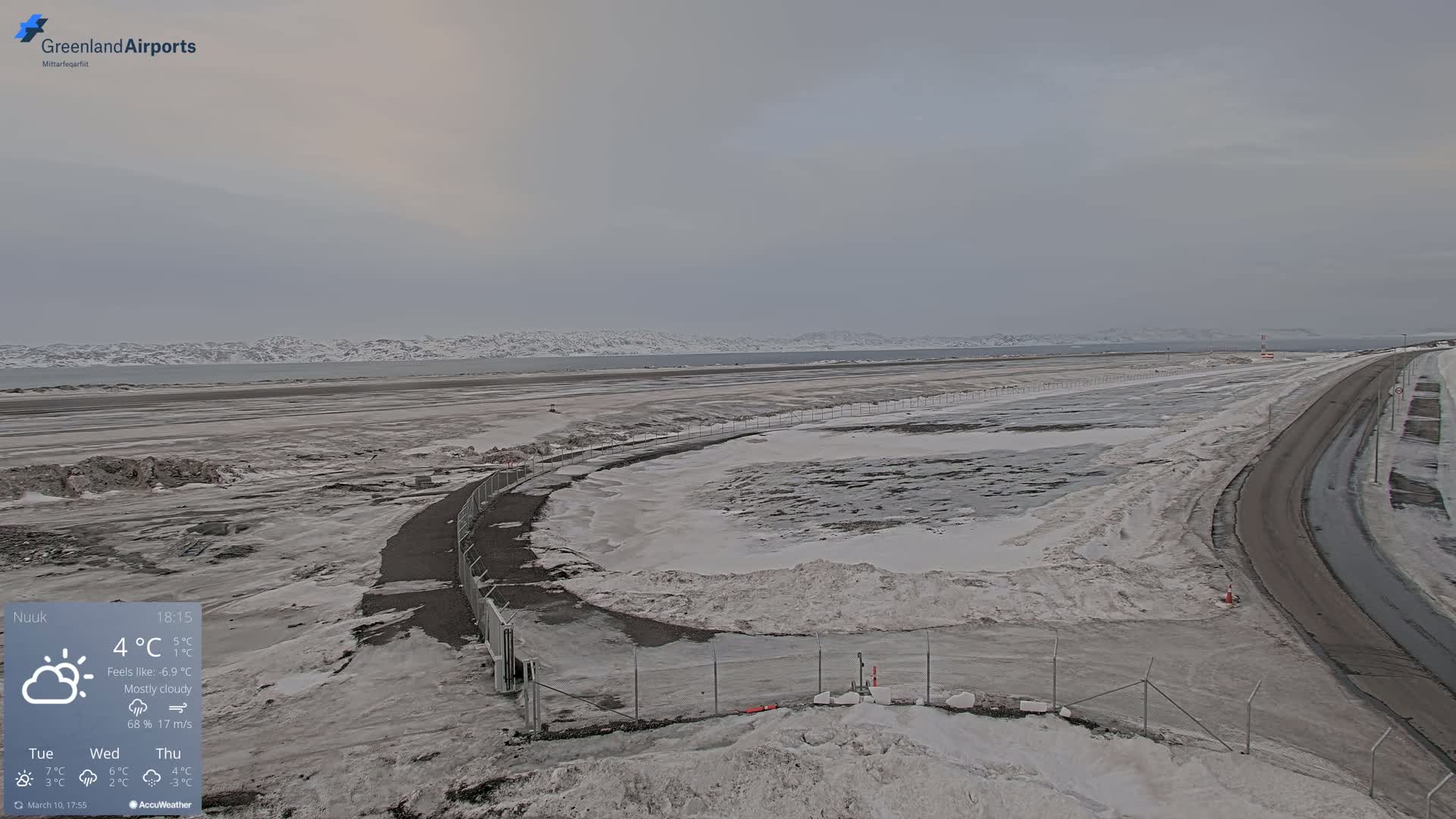 A mostly cloudy, cold day reveals a snow-covered airport tarmac and adjacent road, with distant snow-capped mountains visible on the horizon.