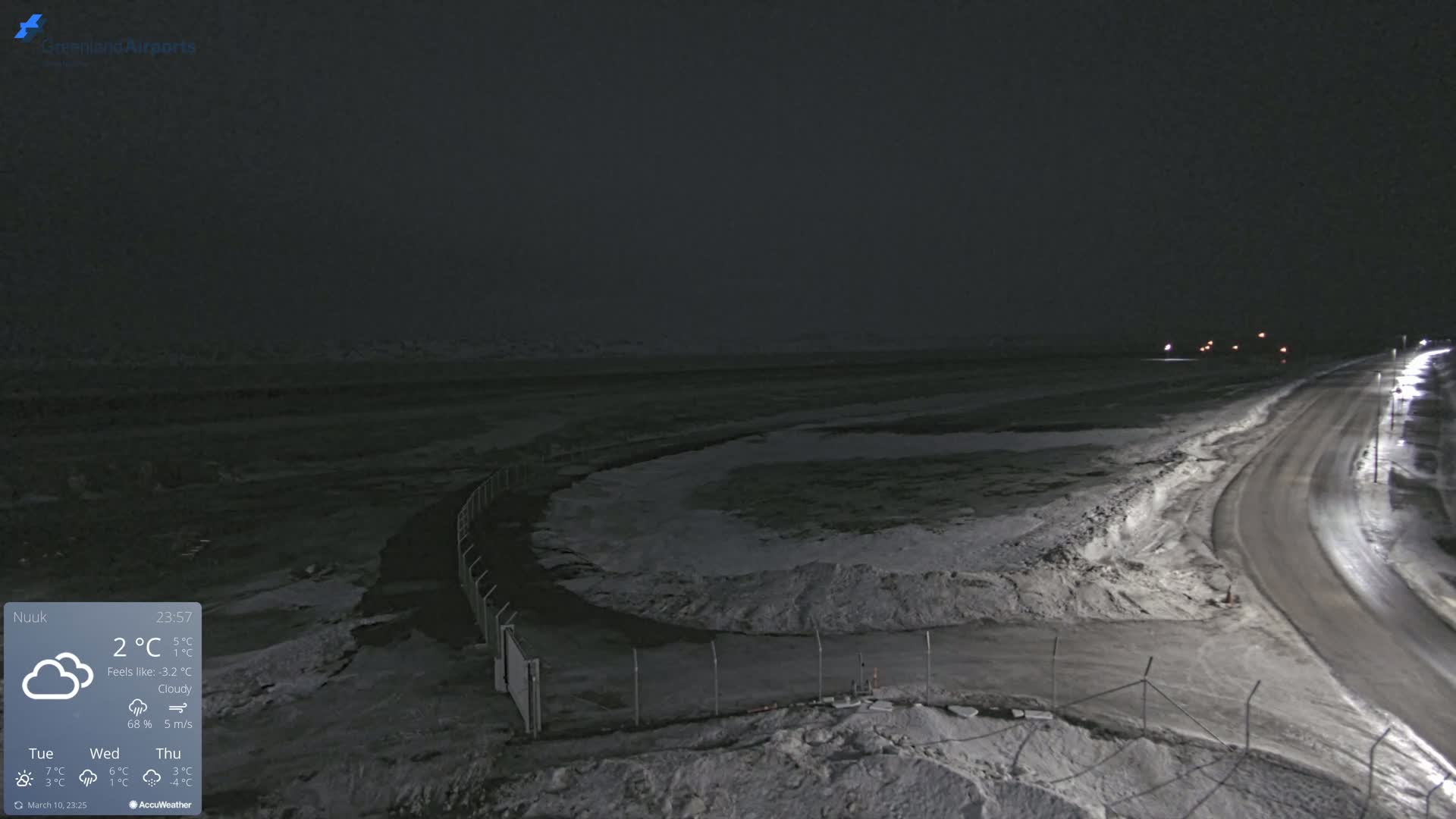 A dark, cloudy night scene shows a snow-covered road curving past a snow-dusted, fenced-in area.