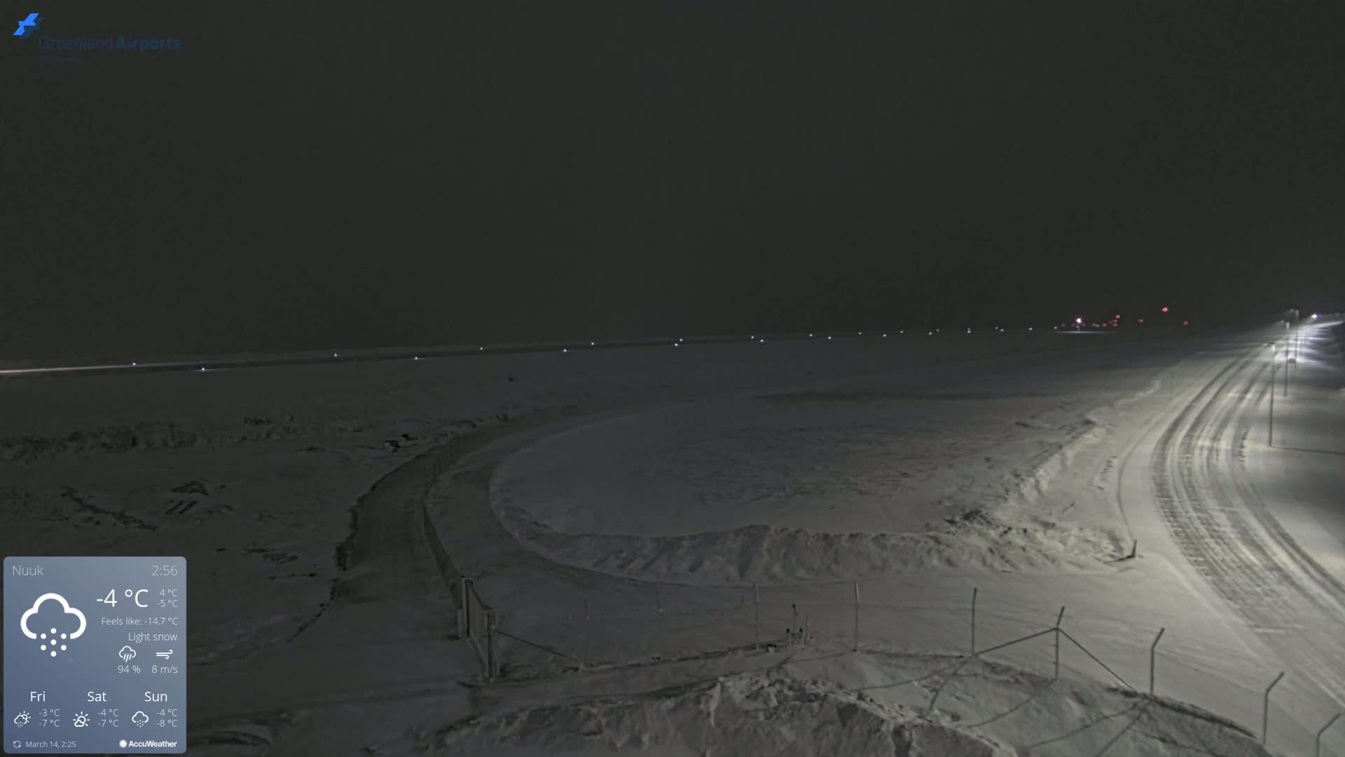 A snow-covered airfield at night under light snow with a road leading away from the scene.