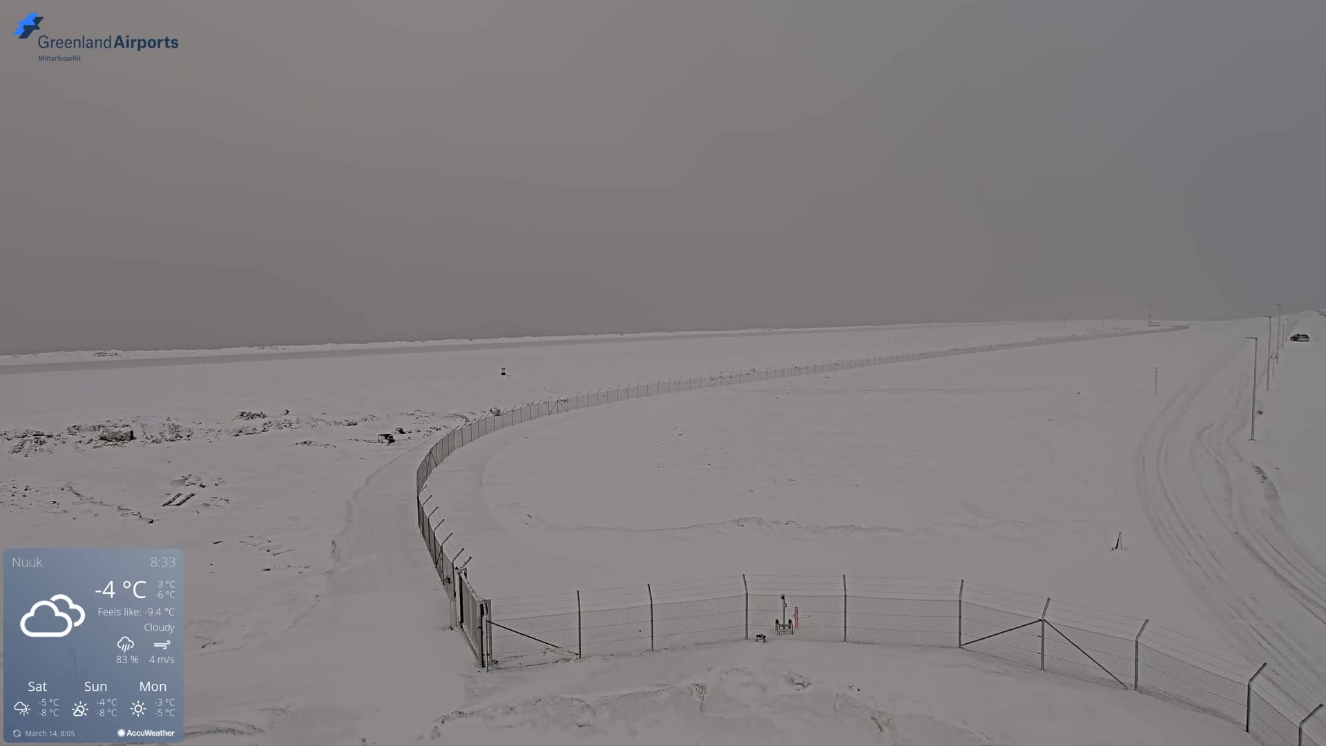 A snow-covered landscape with a chain-link fence and tire tracks in cloudy conditions.