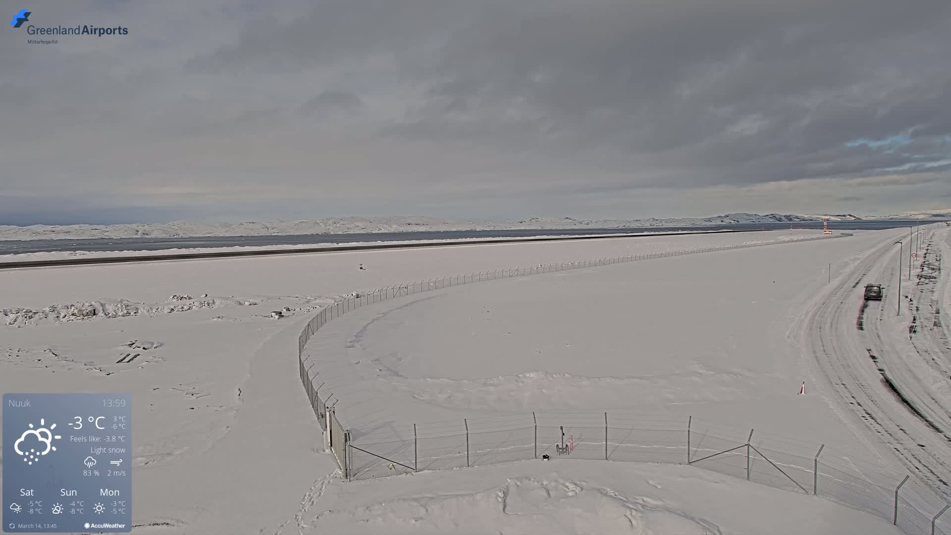 A snow-covered airport runway and surrounding area are visible under a cloudy sky with light snow falling.