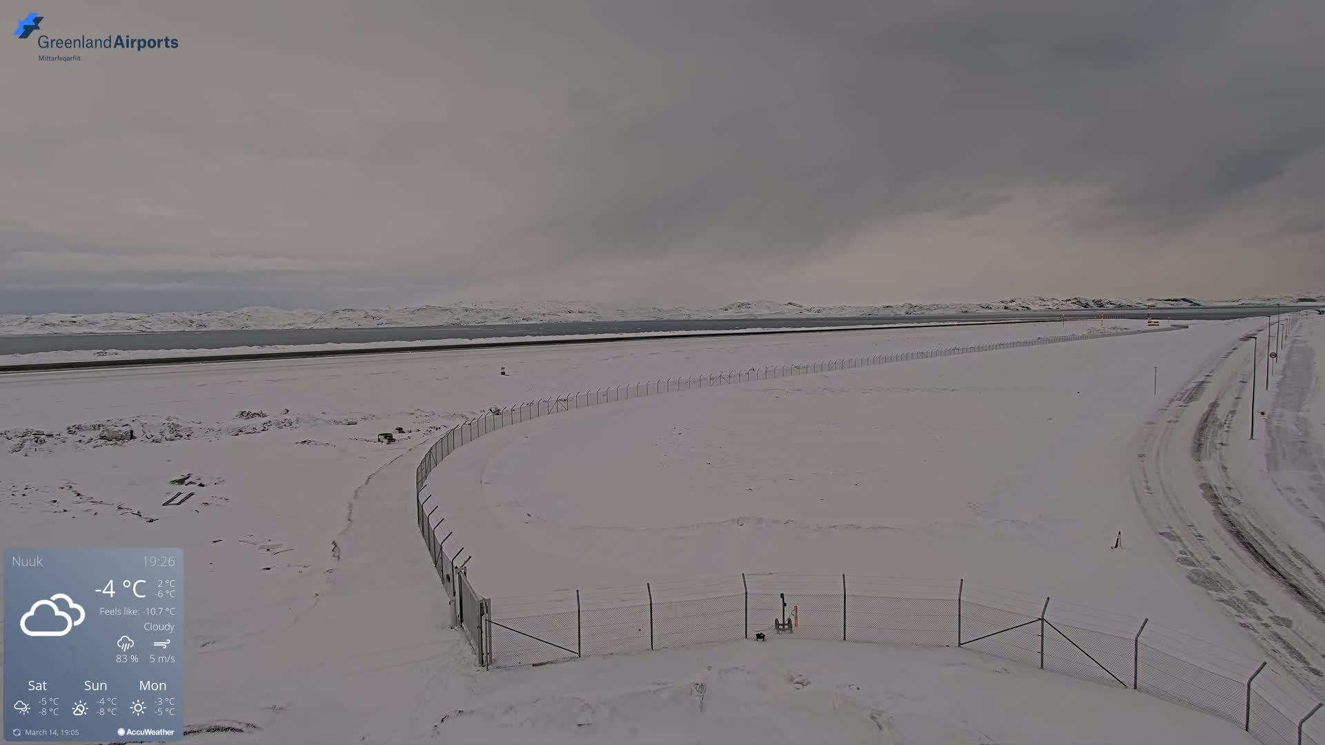 A snow-covered airport tarmac and surrounding area extend to a body of water under a cloudy sky.