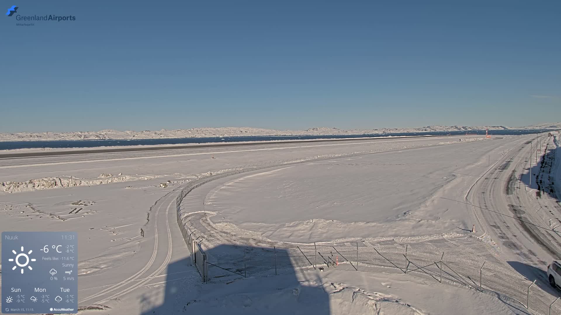 A snow-covered airport runway and access road extend toward a calm body of water and distant mountains under a sunny sky.